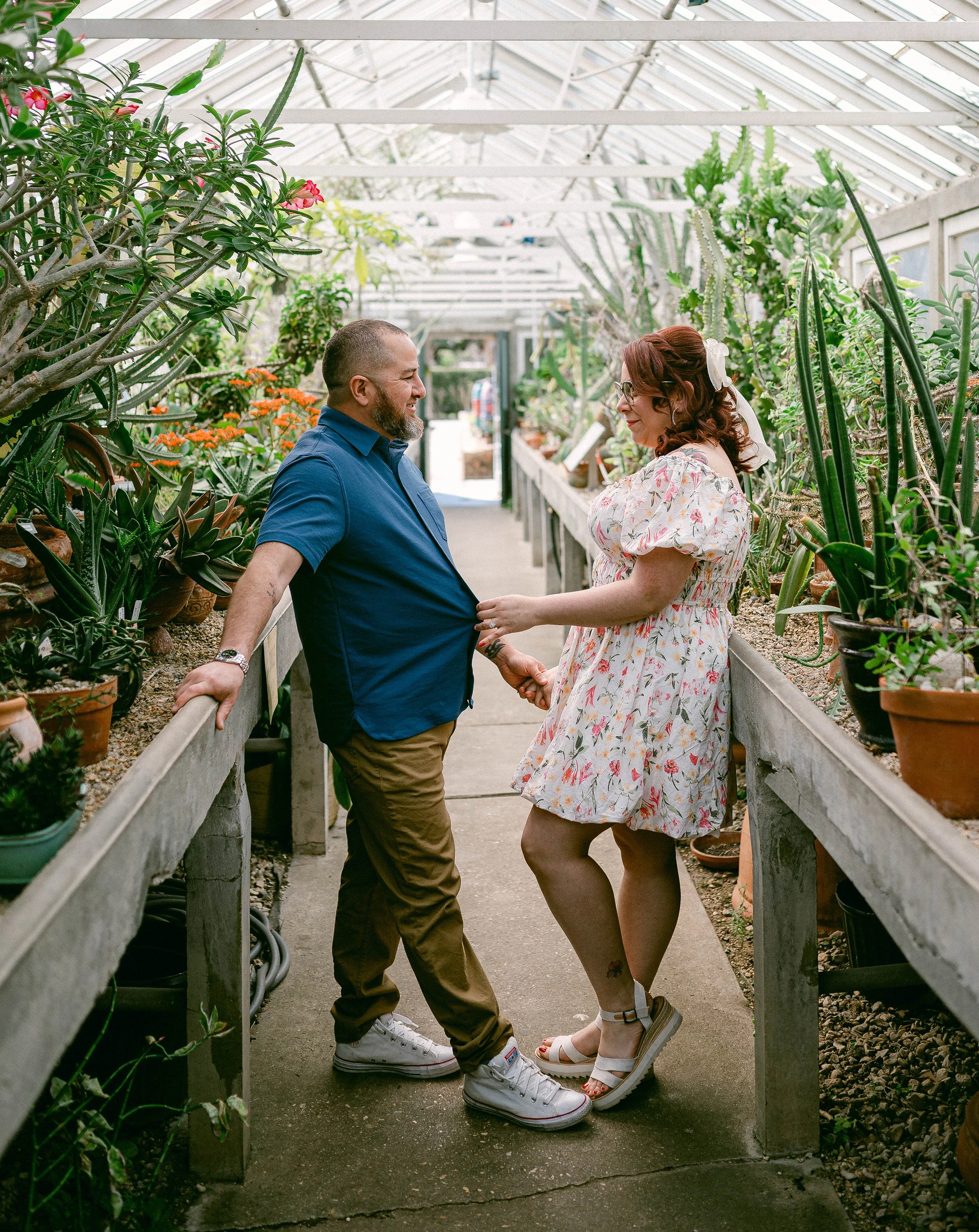 An engaged couple holding hands and smiling at each other inside a greenhouse filled with various plants and flowers.