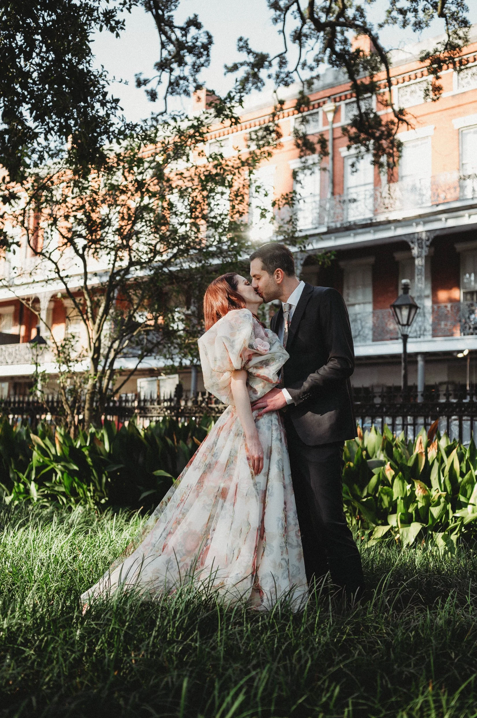 A bride and groom share a kiss outdoors in New Orleans with balconies and lampposts in the background, surrounded by greenery and trees.