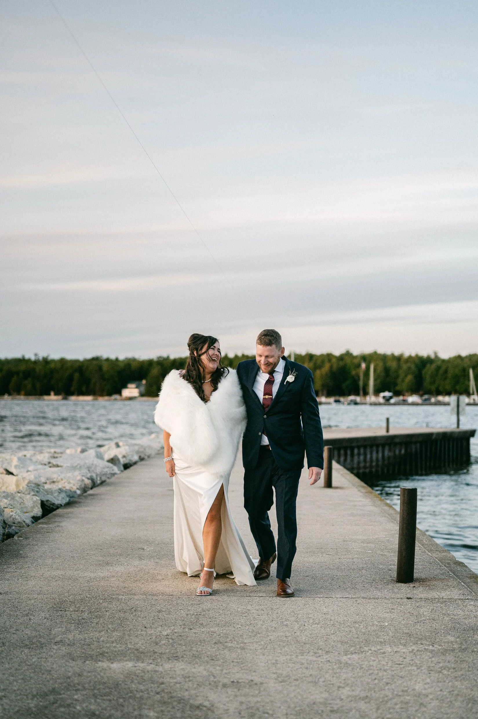 A bride and groom walking together on a pier on Lake Michigan with a tree-lined shoreline in the background, during sunset.