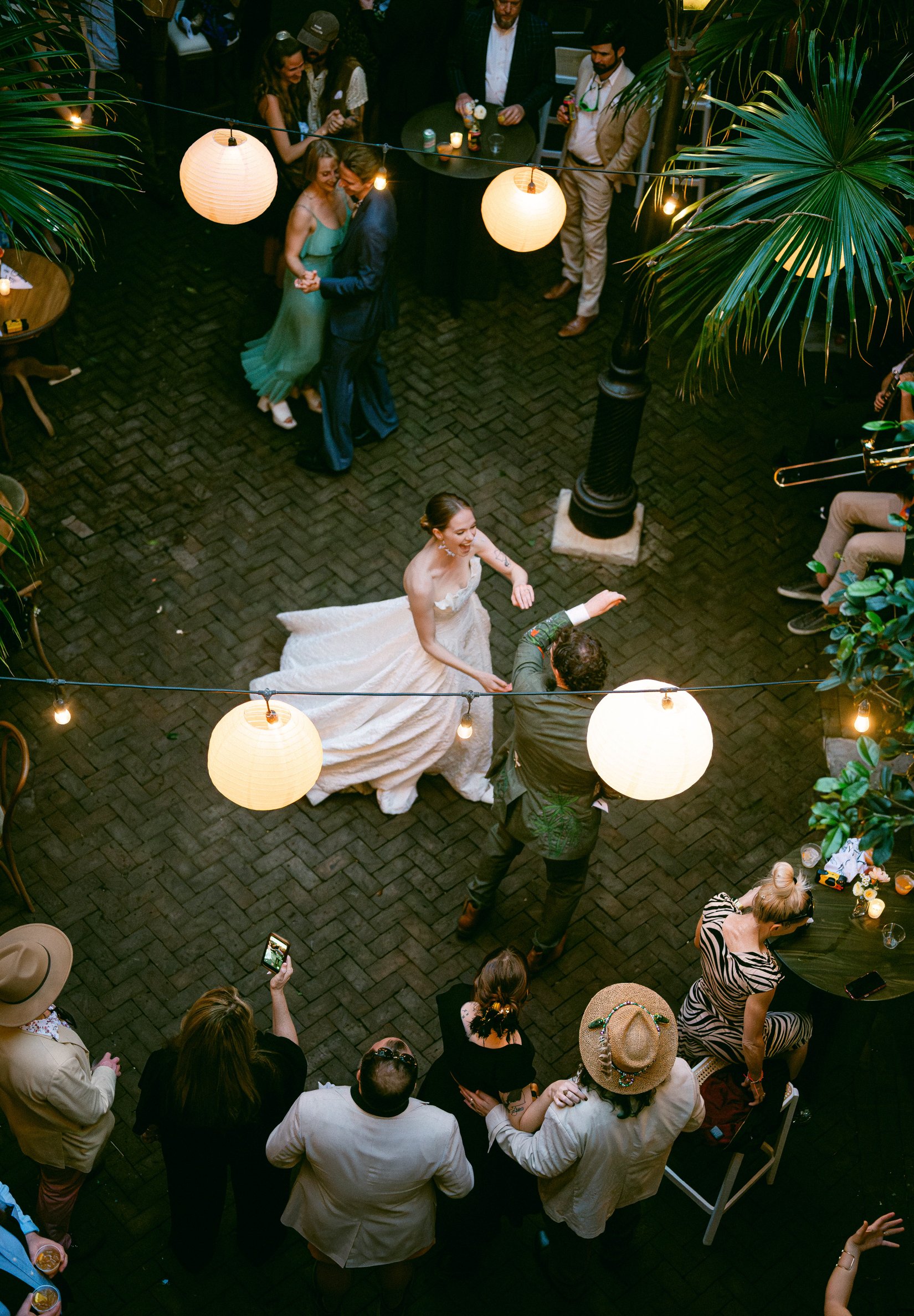 A wedding reception with a bride and groom dancing in the center, surrounded by guests, string lights, and hanging paper lanterns in a New Orleans courtyard.