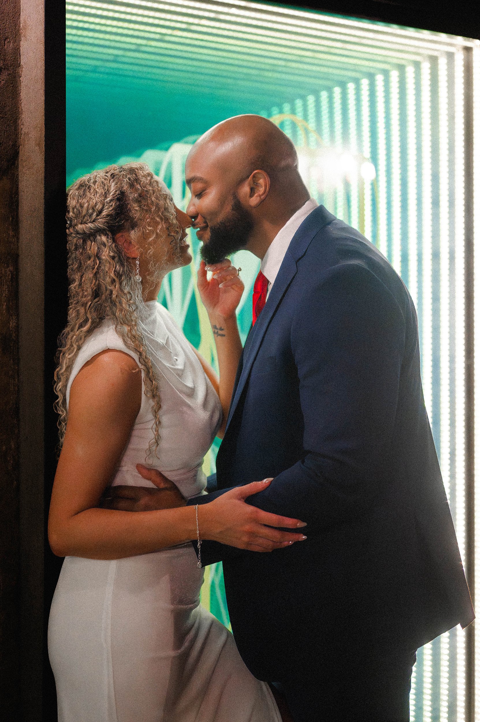 An engaged couple dressed in formal attire, about to kiss, standing close to each other with eyes closed, in a room with a colorful LED light background.