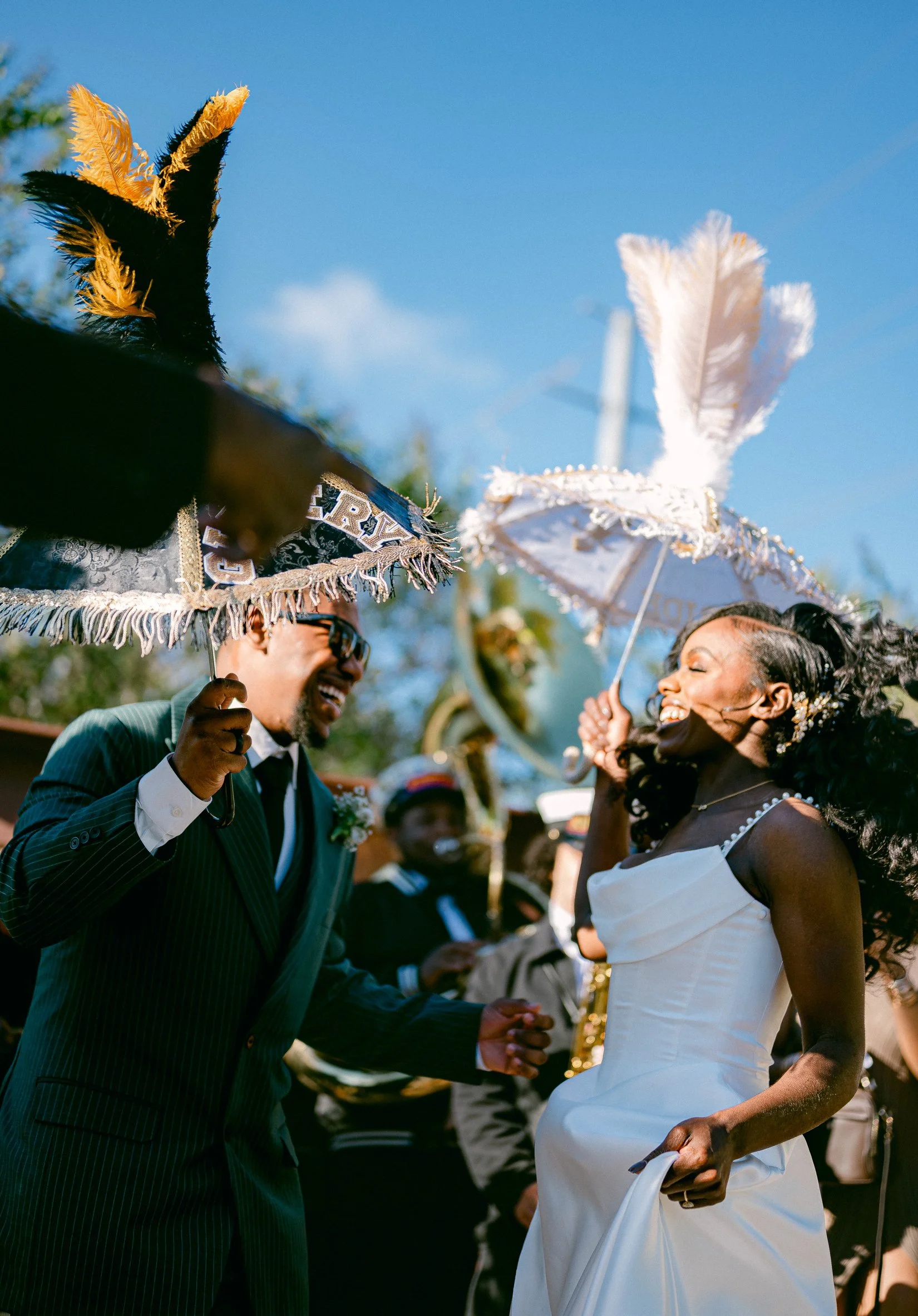 A bride and groom smiling and holding hands during their wedding second line, with guests in the background, under decorative umbrellas, on a sunny day.
