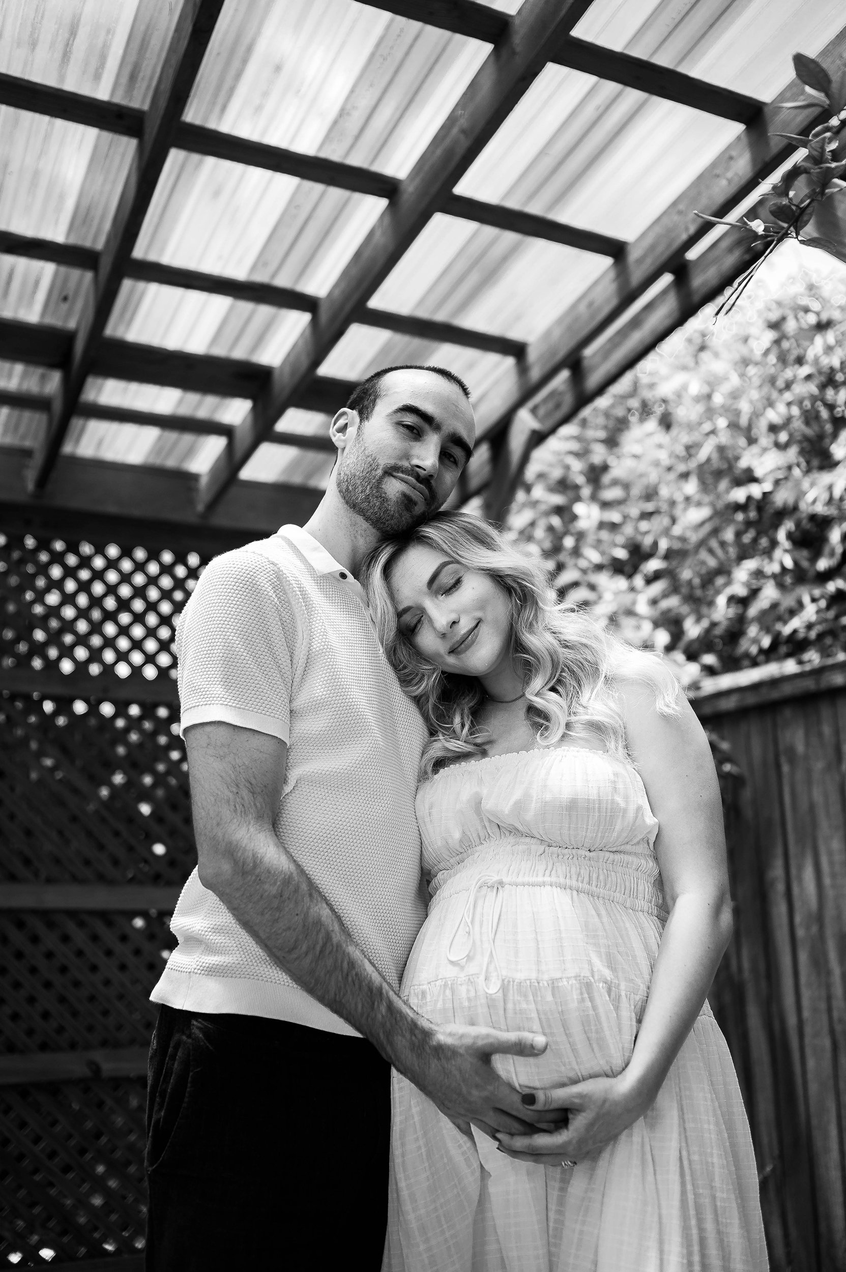 A man and a pregnant woman standing closely together under a wooden pergola, with the woman resting her head on the man's shoulder, all in black and white.