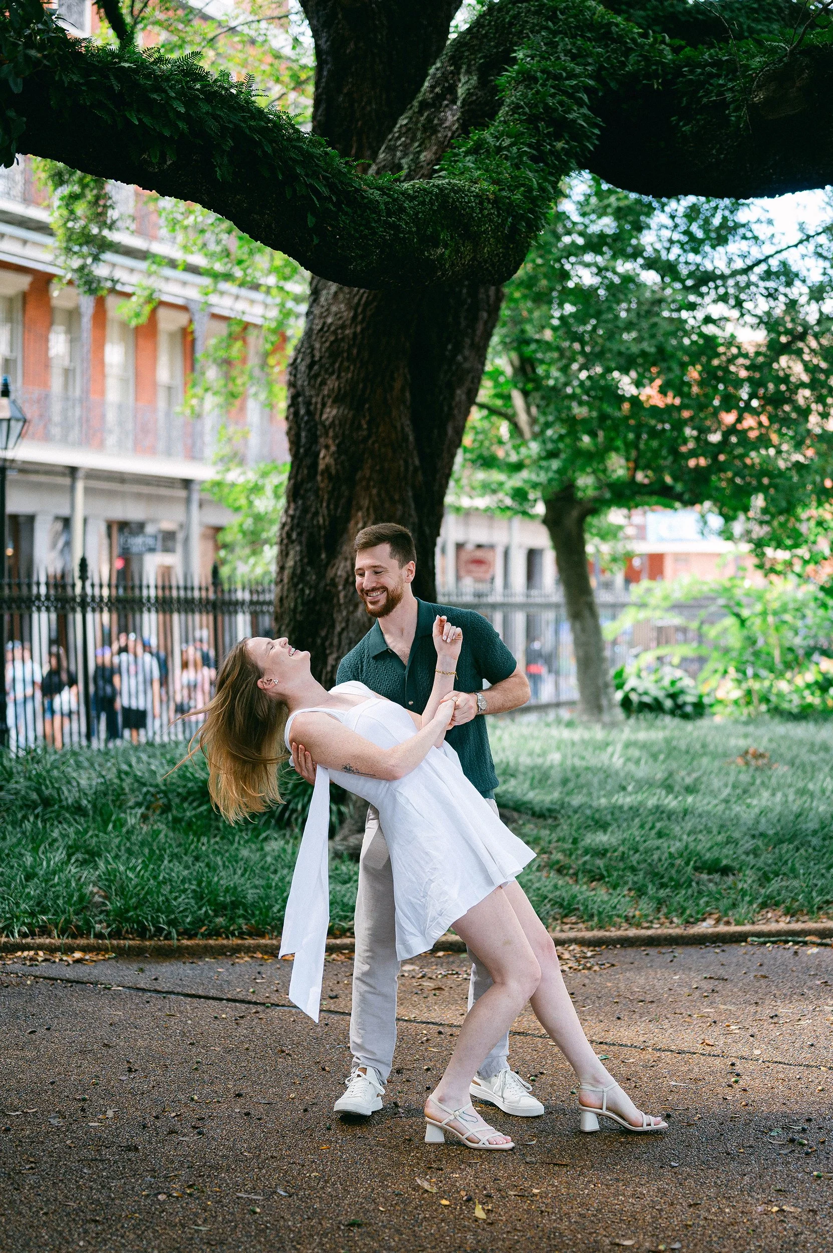 A couple dancing outdoors in a New Orleans park with a large tree and city buildings in the background. The scene is lively and romantic.