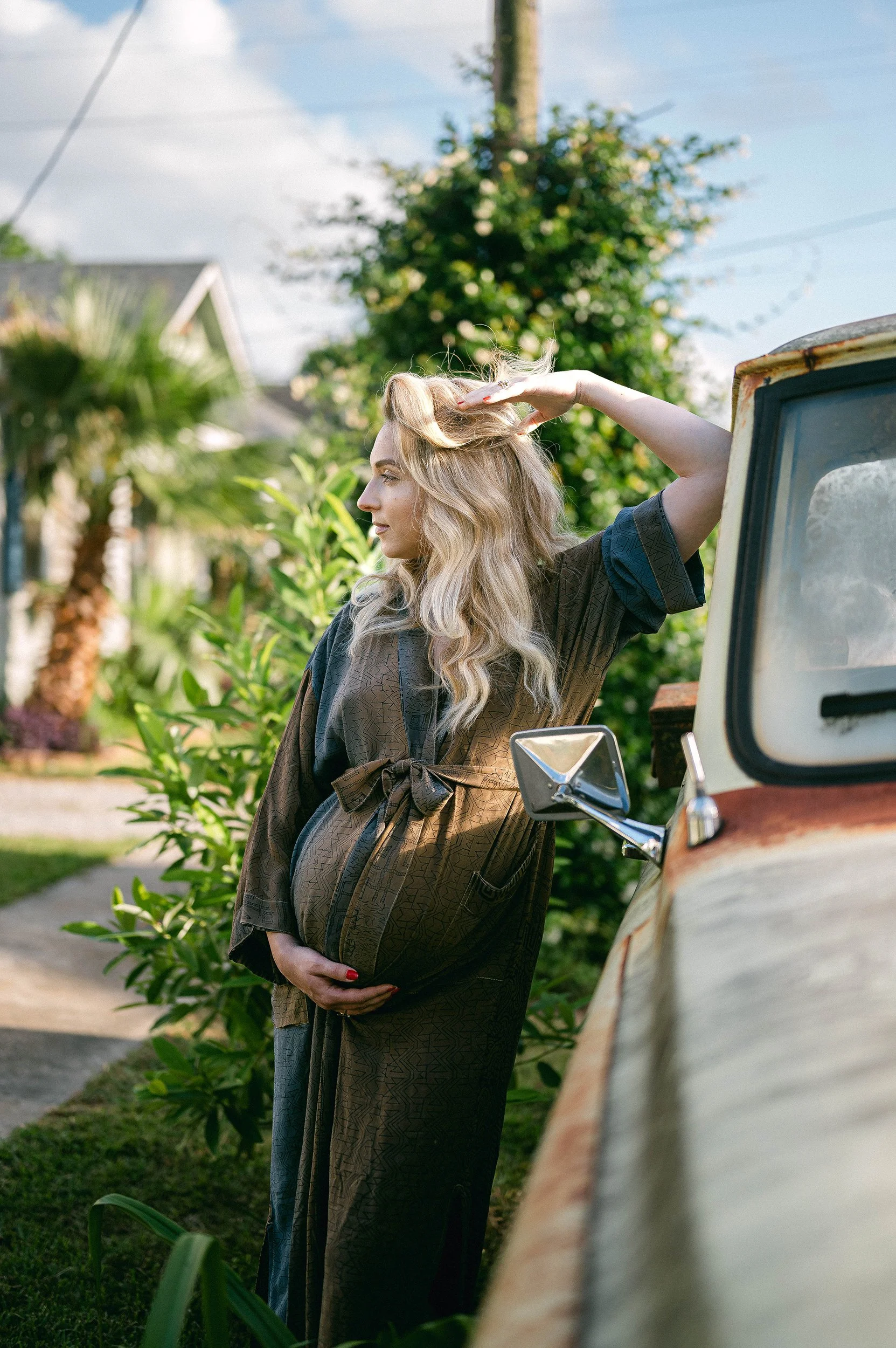 A pregnant woman with long blonde wavy hair in a brown patterned oversized dress, standing beside an old pickup truck with a rusted hood, in a lush garden.
