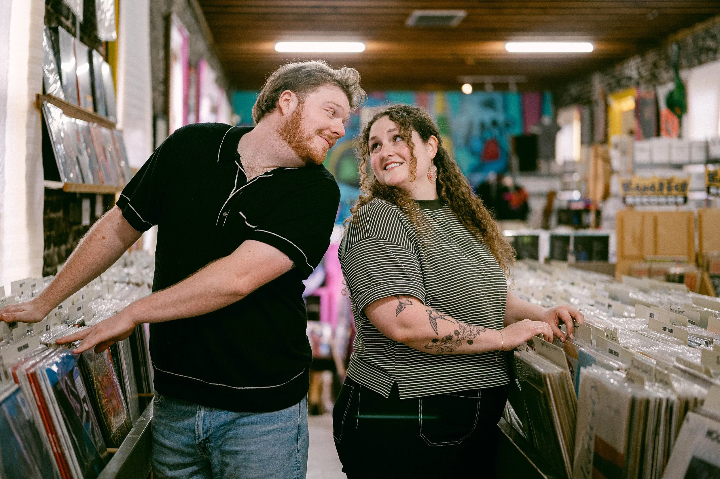 An engaged couple browsing records in a store, smiling at each other, with colorful artwork and shelves of vinyl in the background.