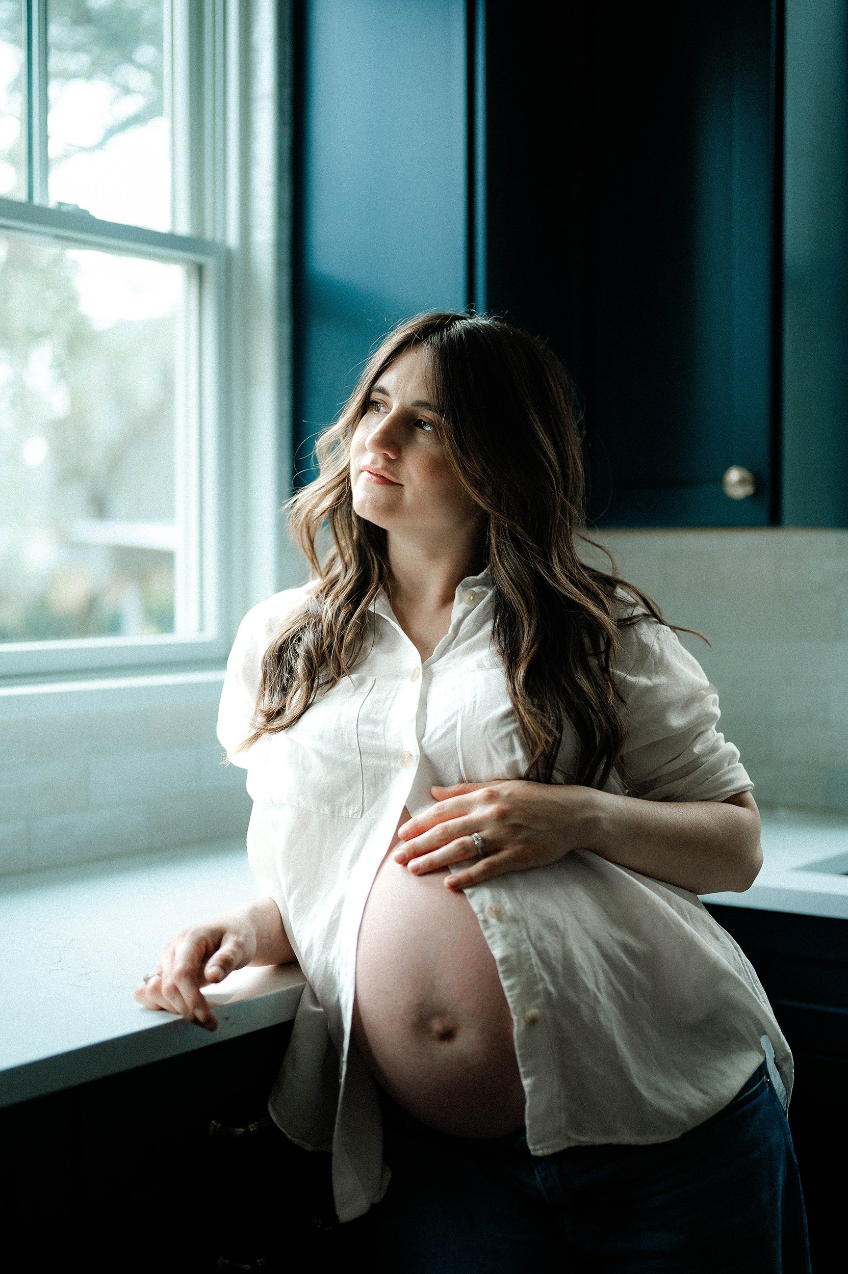 A pregnant woman with long wavy hair and a white shirt sitting by a window with natural light, looking thoughtfully outside.