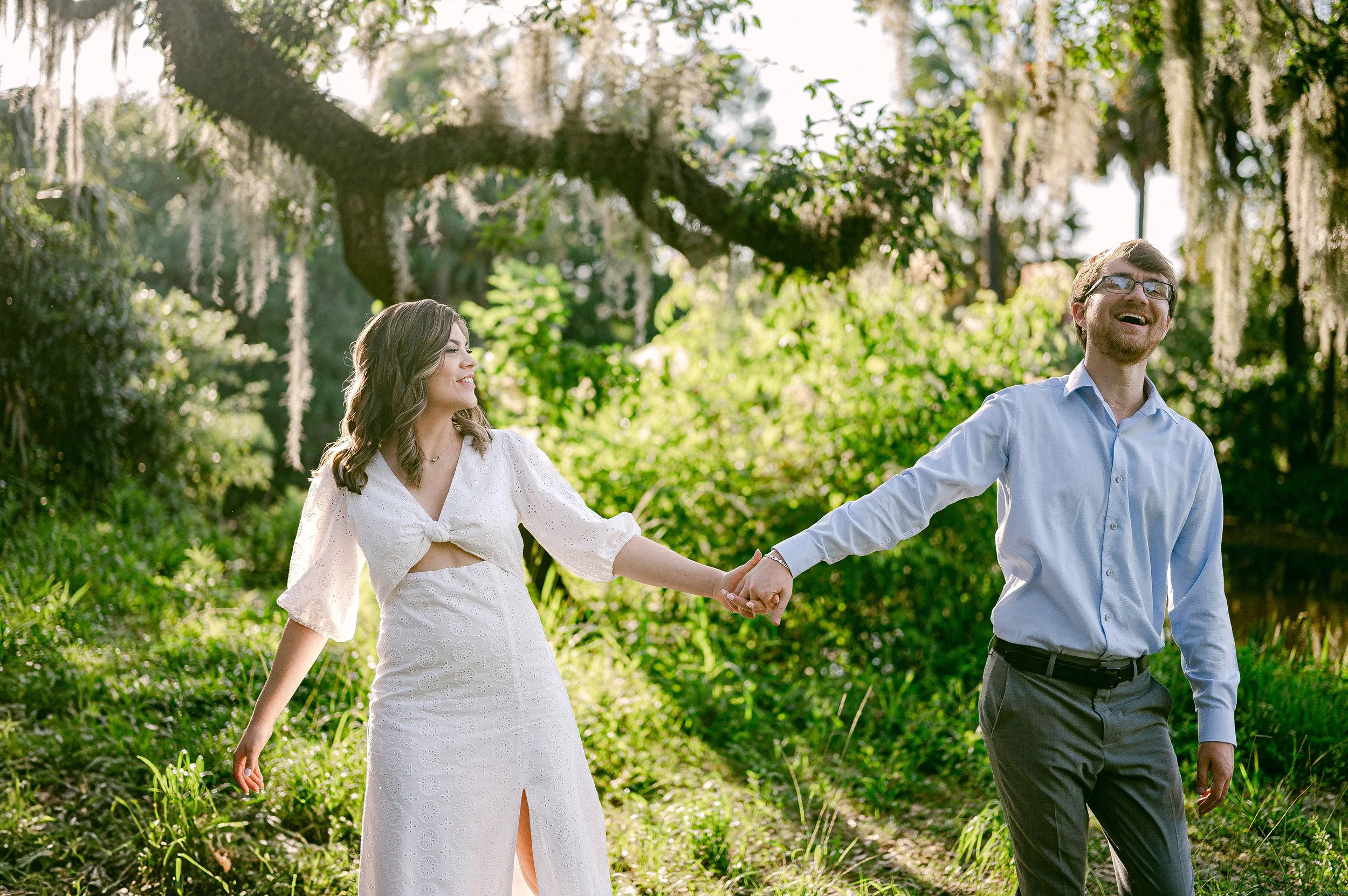 A smiling engaged couple holding hands and walking through a lush green forest in the daytime, with sunlight filtering through the trees.