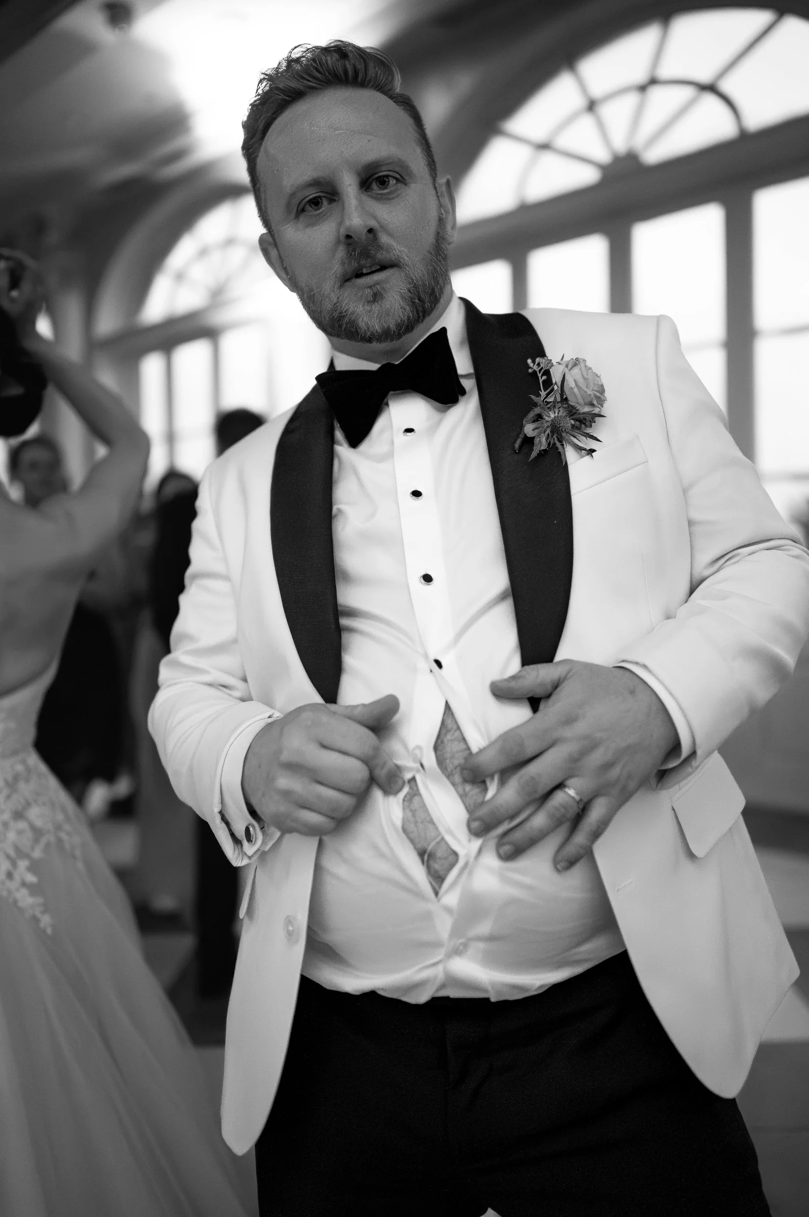 Black and white photo of a groom in a white tuxedo with a boutonniere, opening his shirt to show a giant rip down the front.