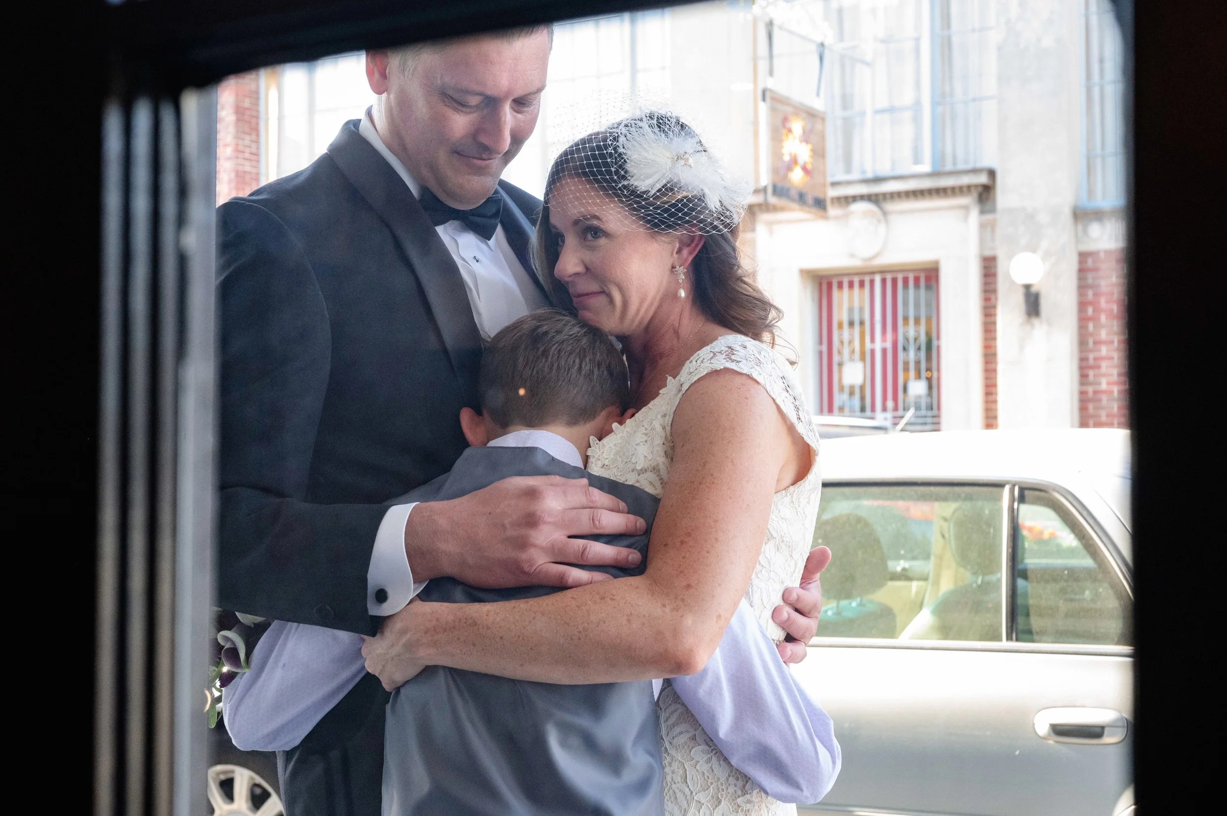 A bride and groom and their young son embrace in a hug following a wedding ceremony, as seen through a glass window.