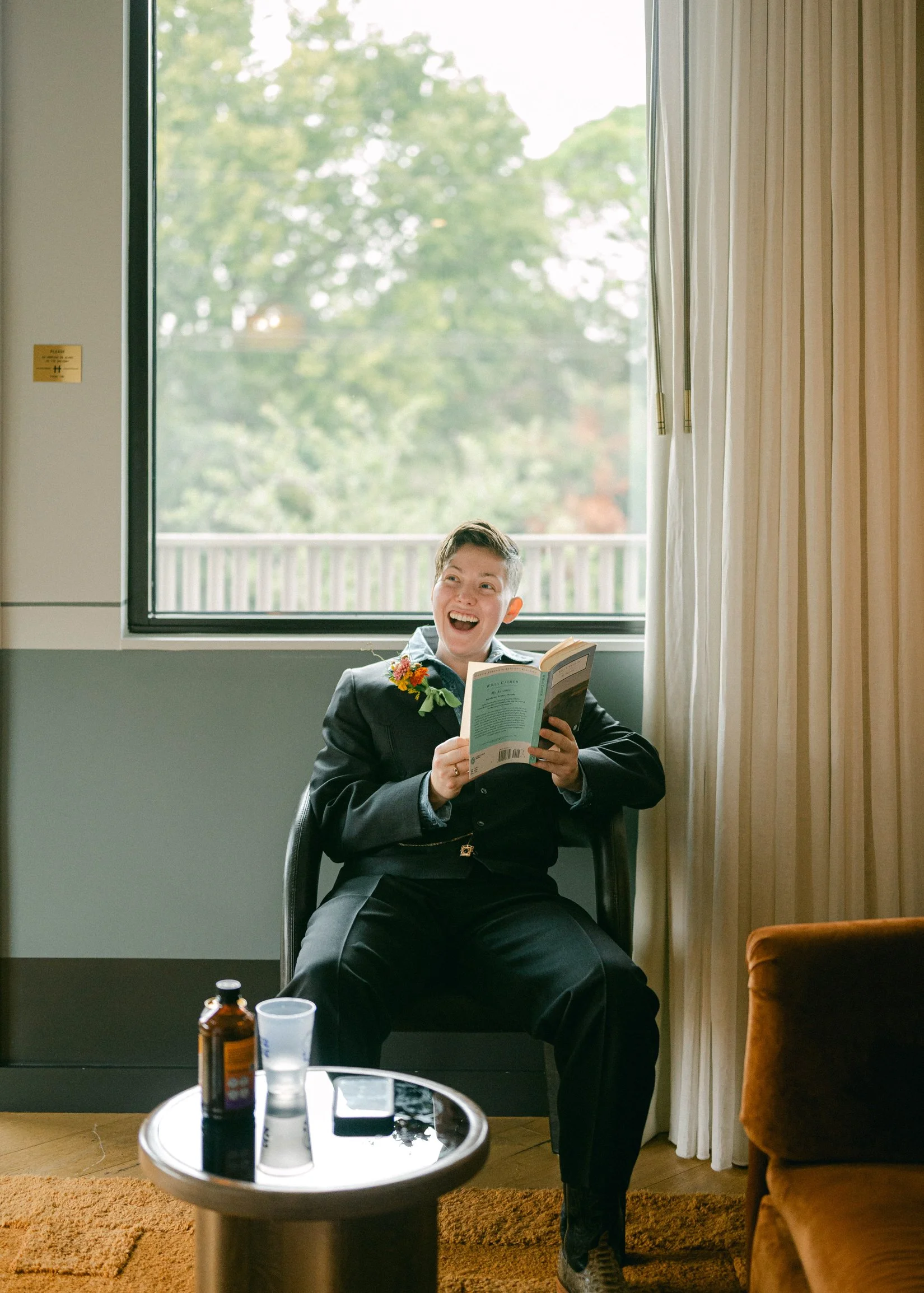 A newlywed, wearing a dark blazer, sitting on a chair by a window, reading a book and smiling.