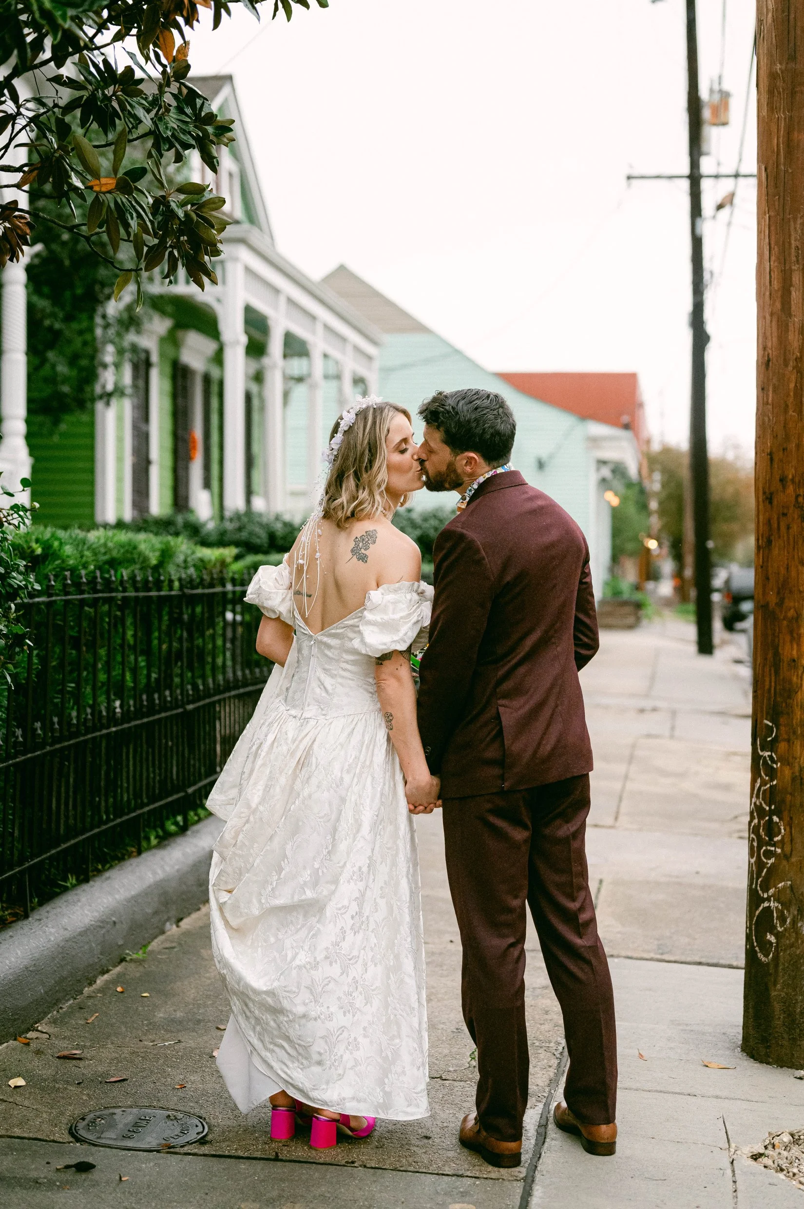 A couple in wedding attire is kissing on a sidewalk in a New Orleans neighborhood with colorful houses and utility poles in the background.