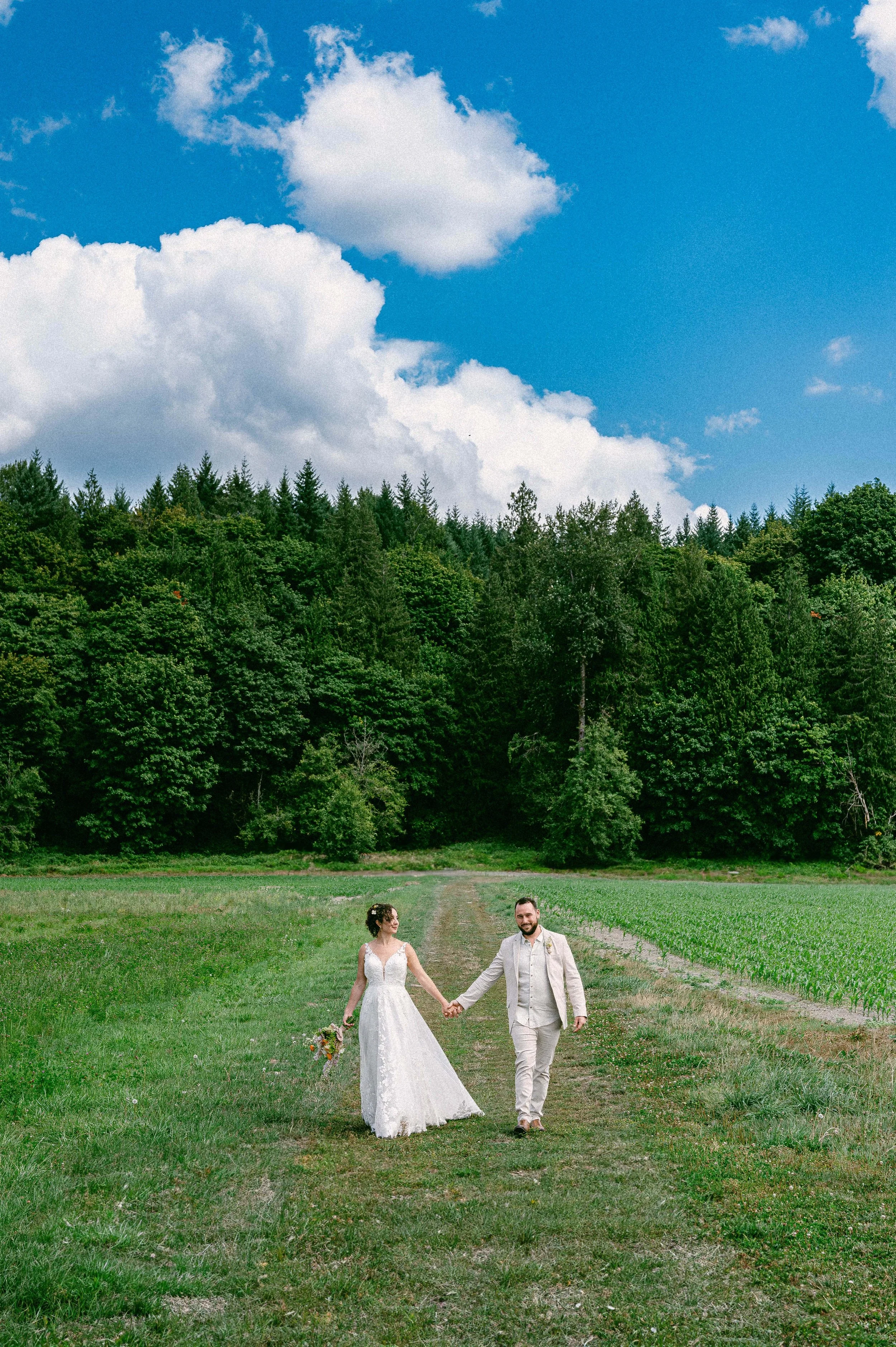 A bride and groom walking hand in hand on a dirt path through a lush green field in Snohomish, Washington, with a dense forest of trees in the background and a partly cloudy blue sky above.