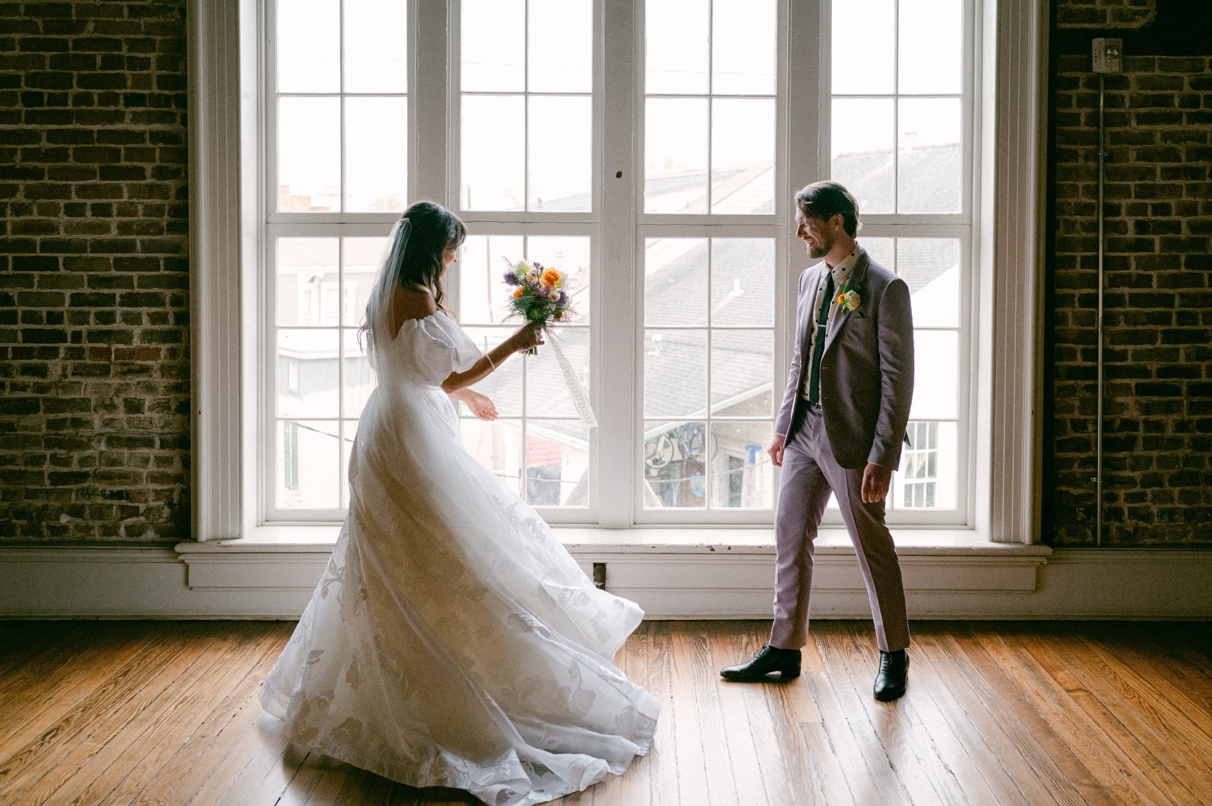 A bride in a white wedding dress holding a bouquet of flowers and smiling at a groom in a lavender suit during their first look.
