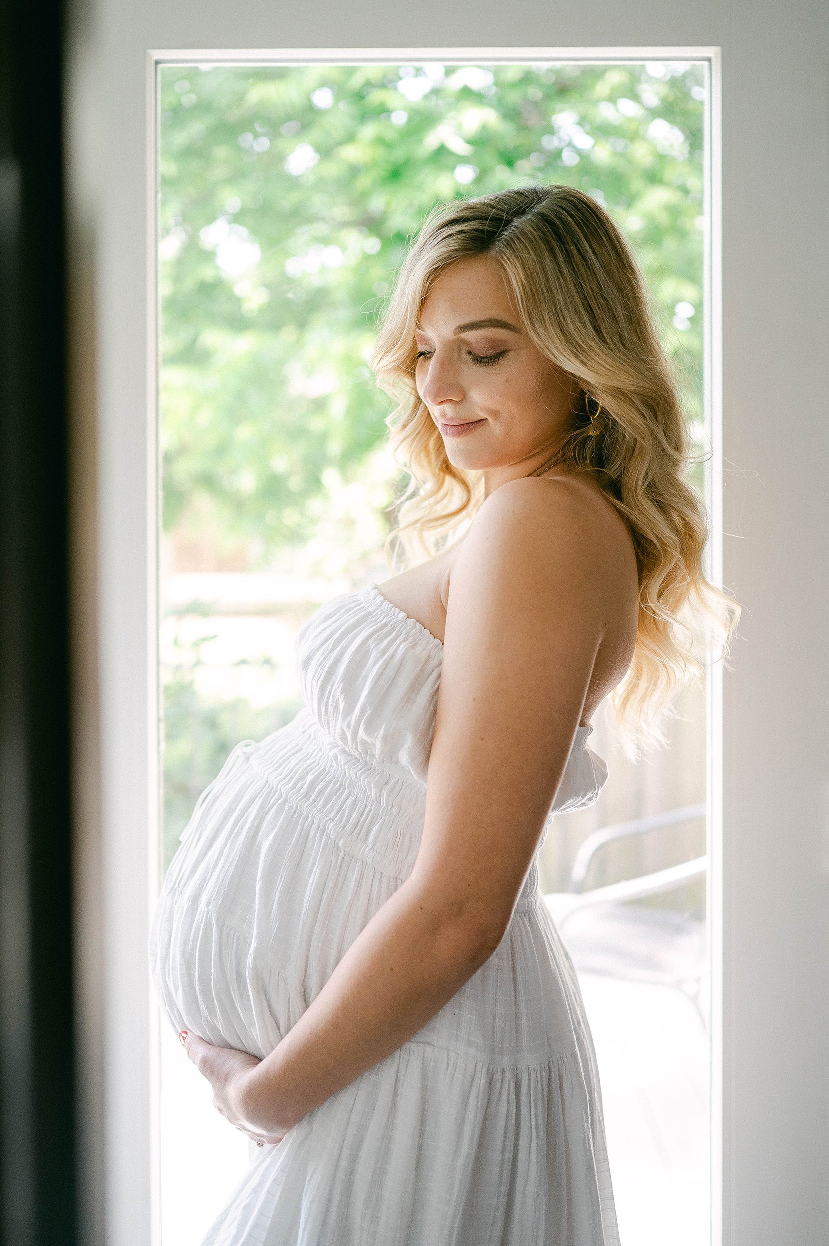 A pregnant woman with wavy blonde hair smiling and gently holding her belly in front of a window with green trees outside.