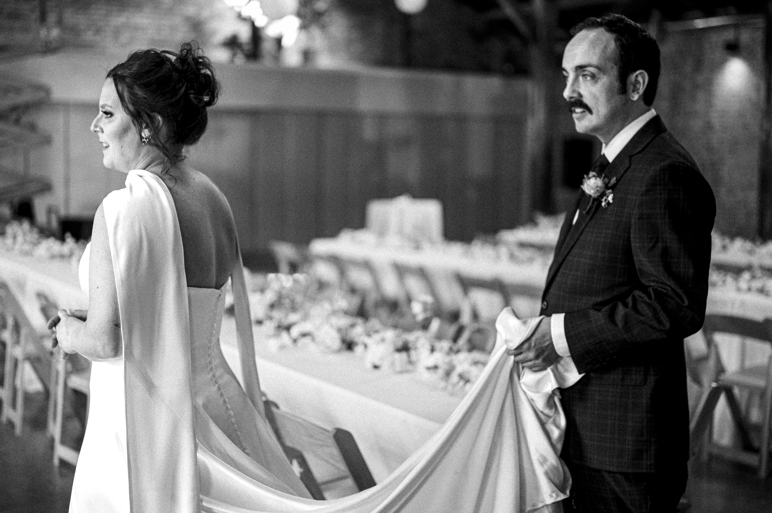 Black and white image of a bride in a wedding dress and a groom in a tuxedo holding his bride's train.