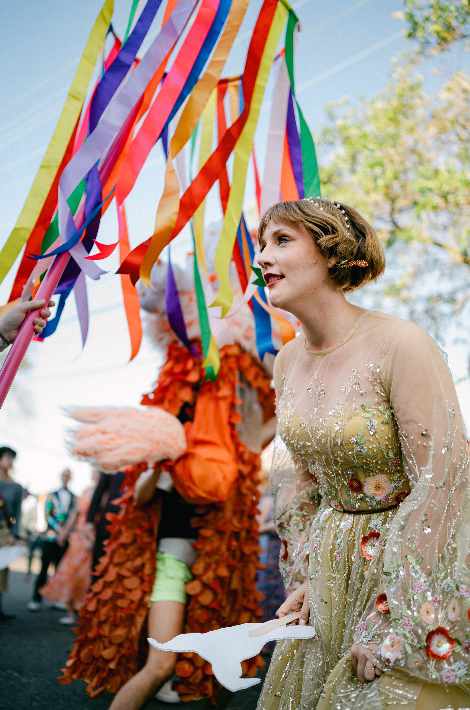 A bride in an ornate, floral-embellished gold wedding gown dances outdoors with other guests in a festive, outdoor setting with trees and clear sky.
