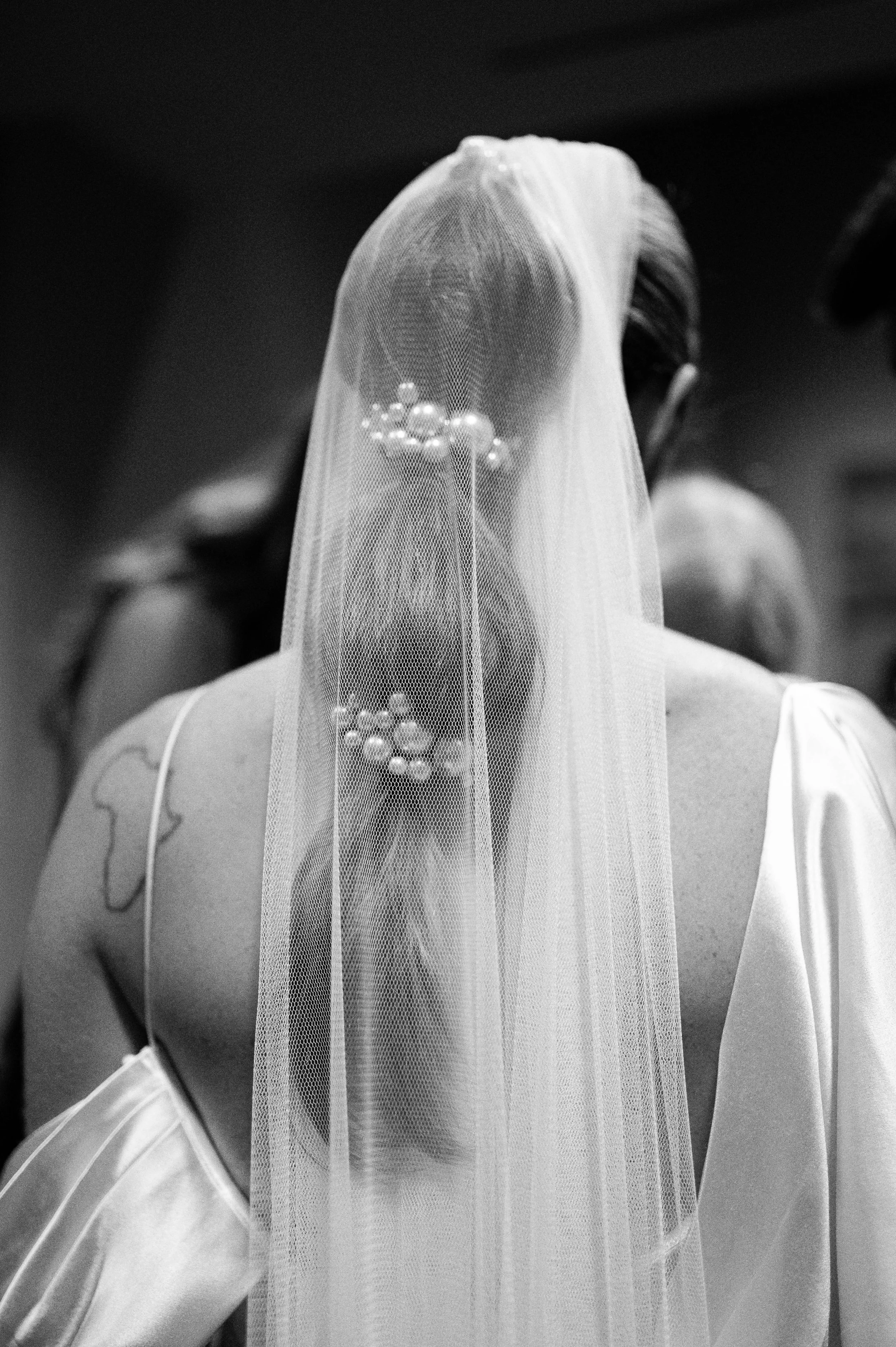 A bride is seen from behind, wearing a wedding veil and a white satin dress, with tattoos visible on their arm.