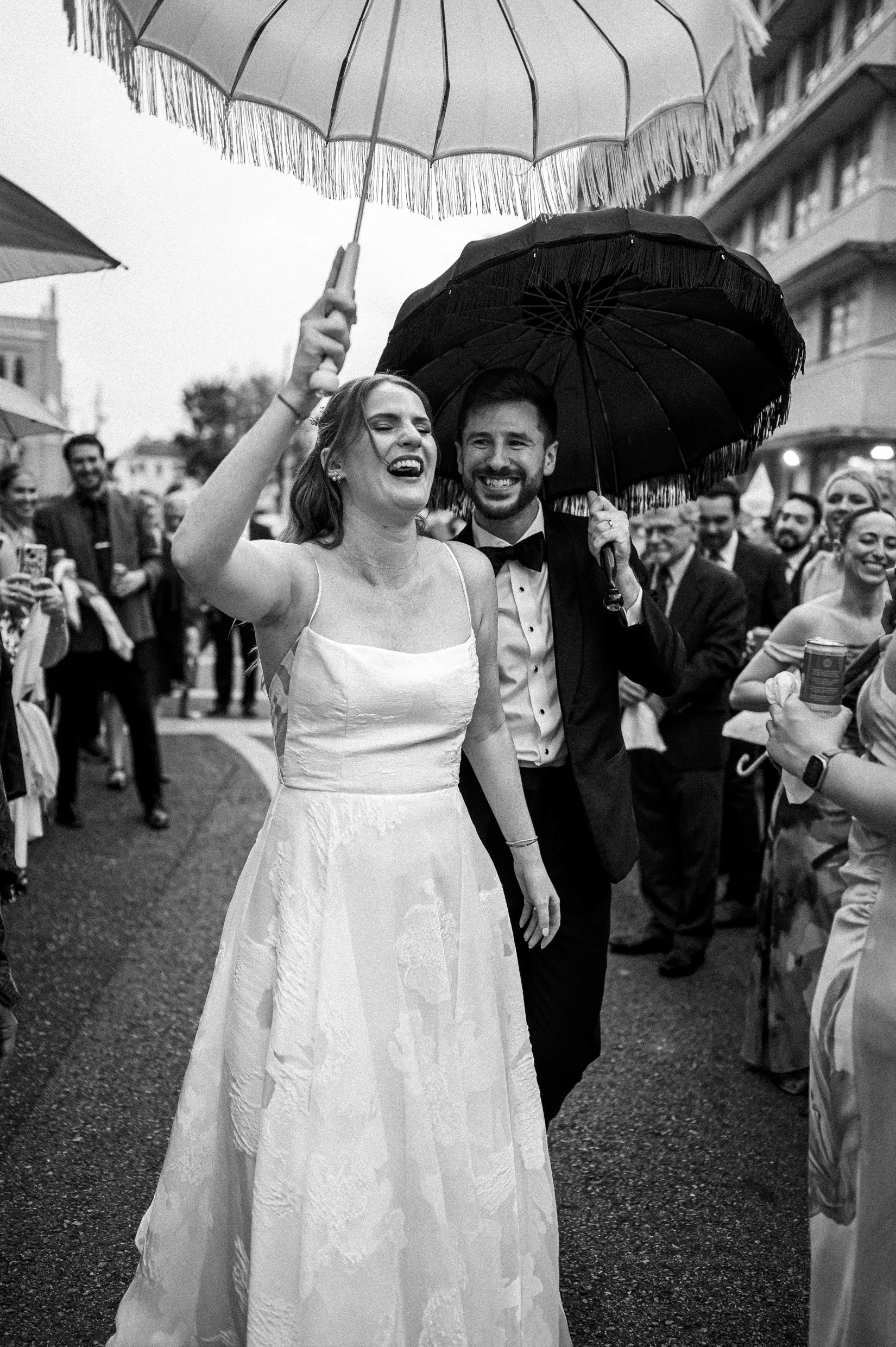 A bride and groom at their wedding celebration, smiling and holding umbrellas, with guests in the background.
