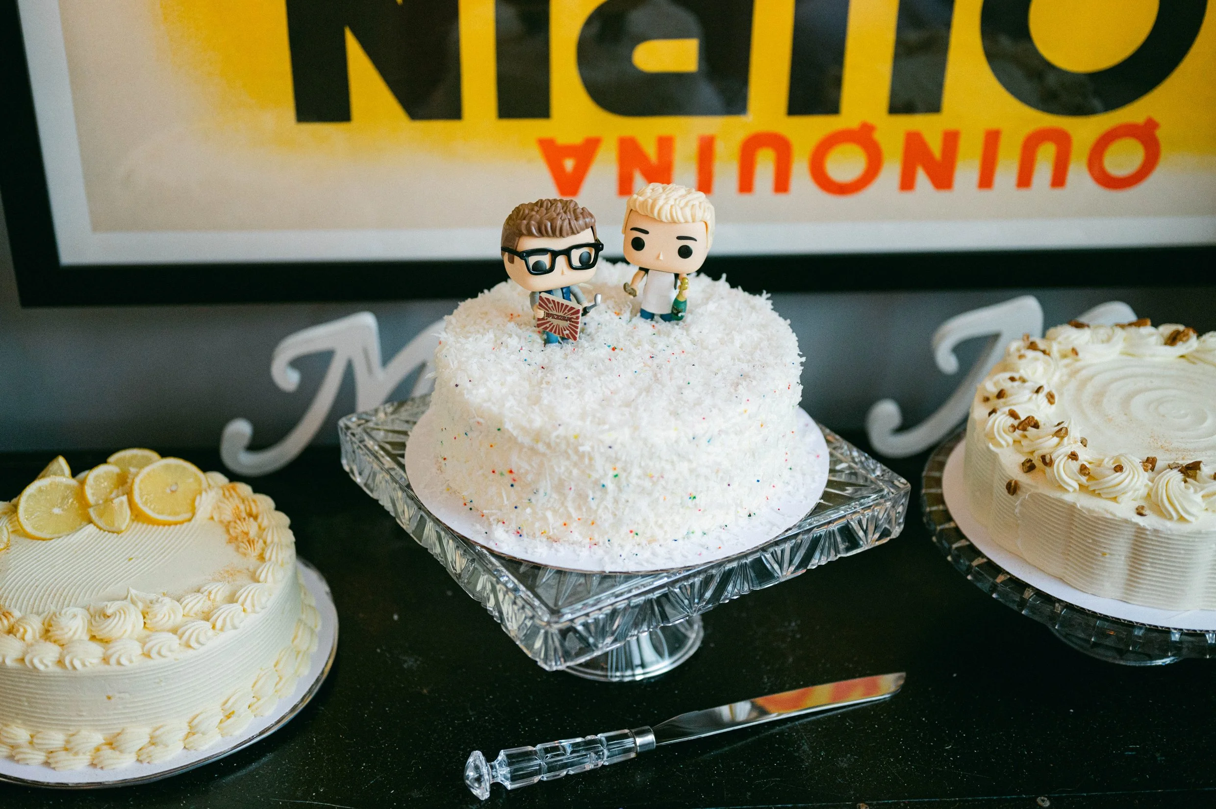 Three wedding cakes, one with coconut flakes and colorful sprinkles topped with figurines of two grooms. 