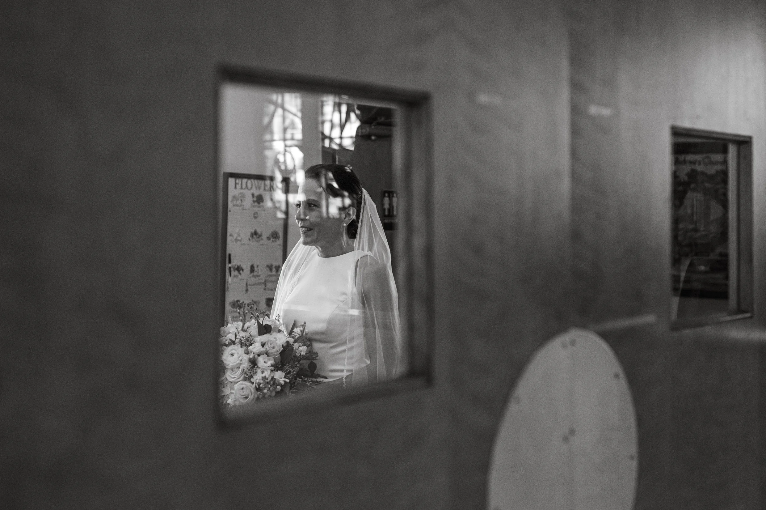 A bride in wedding dress and veil holding a bouquet of flowers, seen through a small window in a church interior.
