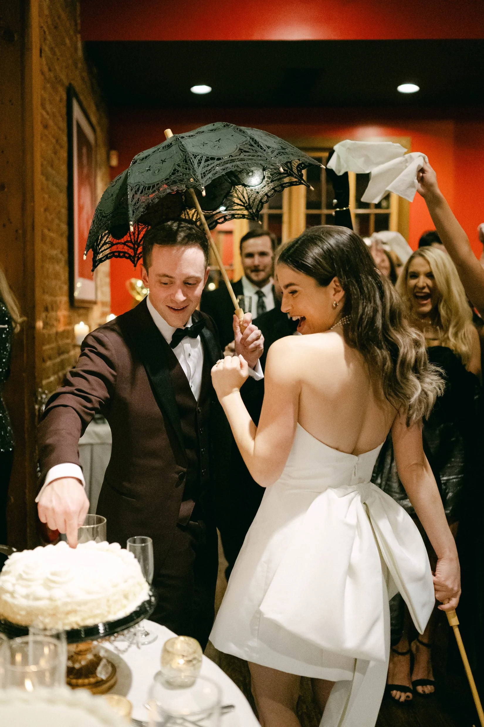 Groom and bride at a wedding reception, the groom holding a black lace parasol, and the bride wearing a white strapless wedding dress with a bow on the back, laughing and eating the cake.