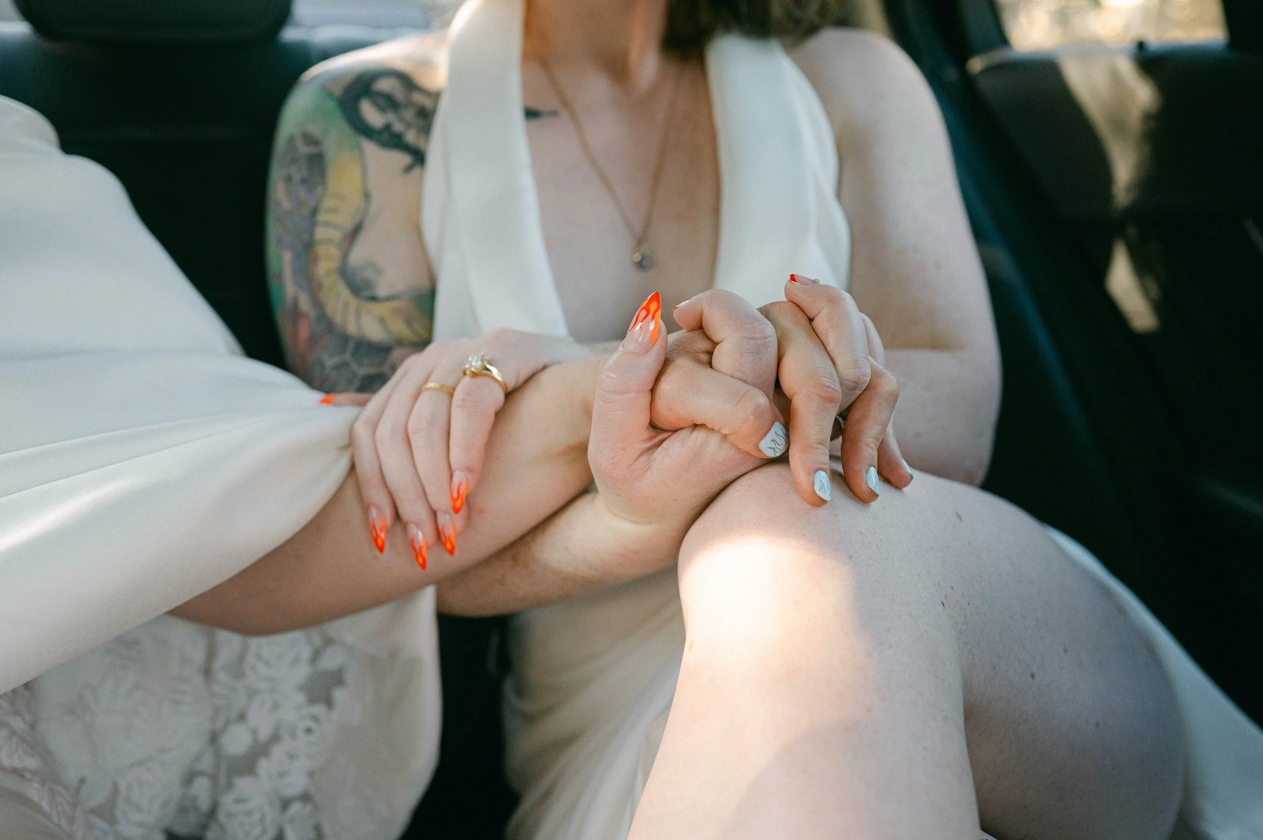 Close-up of two women holding hands, one with colorful nail polish and rings, and the other with a tattoo on her arm, inside a car.