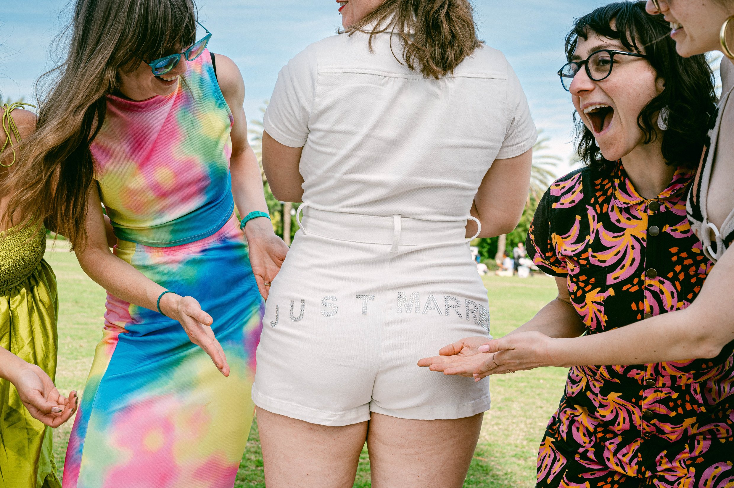 A group of women holding hands and smiling outdoors during daytime, with one woman wearing colorful tie-dye clothing and another in shorts with the words 'Just Married' on it.