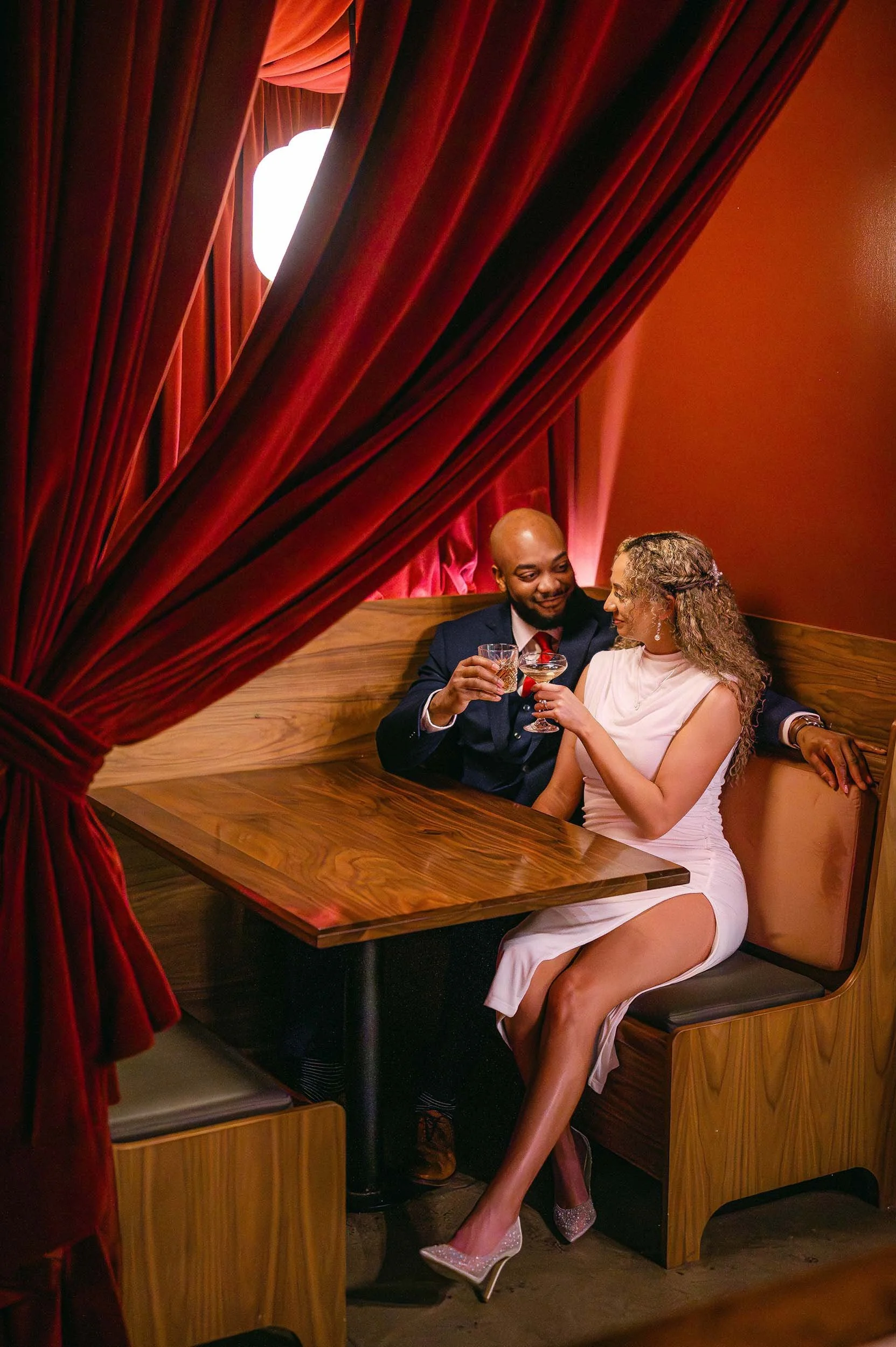 An engaged couple sitting closely in a booth at a restaurant, raising glasses for a toast, with red curtains framing the scene.