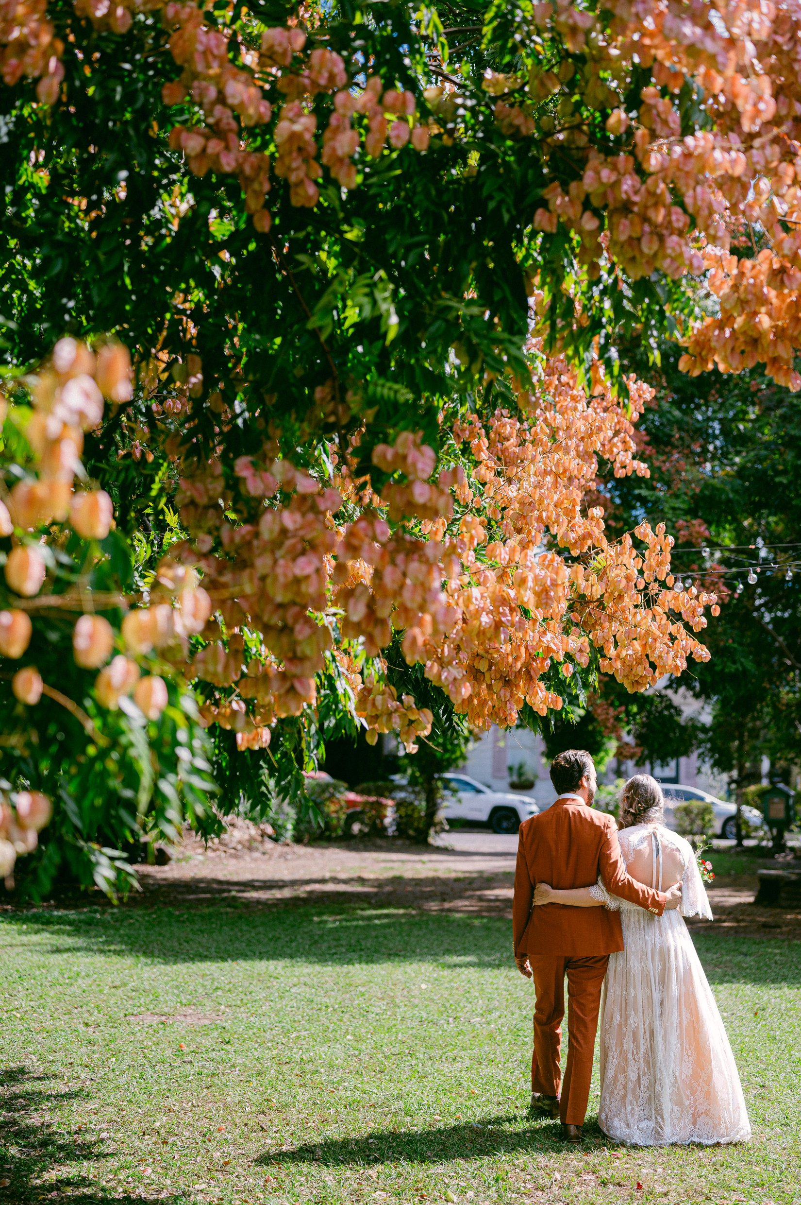 A bride and groom walking together away from their wedding ceremony in a park, surrounded by pink and peach flowering trees.