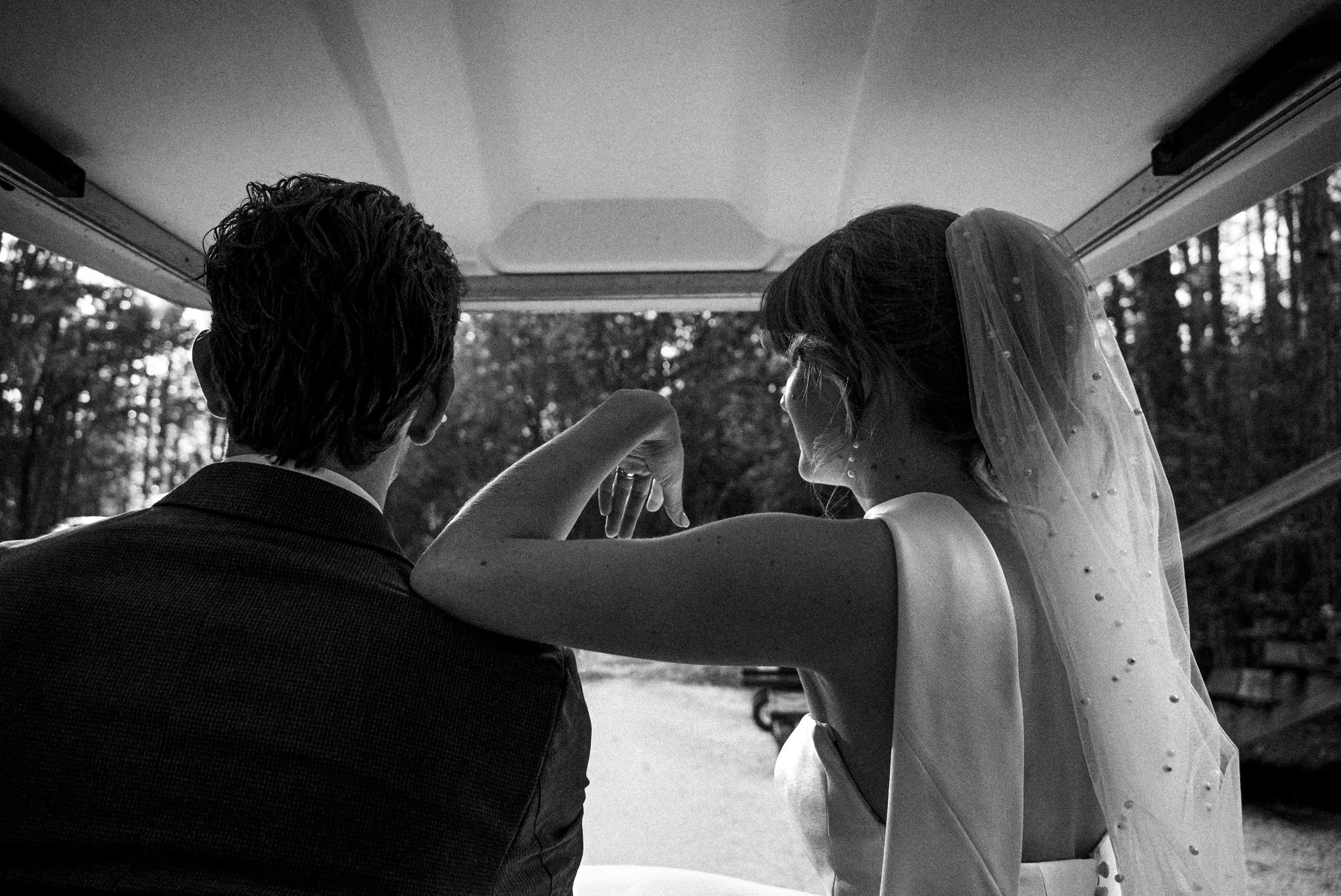 A black and white photo of a bride and groom viewed from behind, sitting in the trunk of a car, holding hands with the bride's veil and dress visible.
