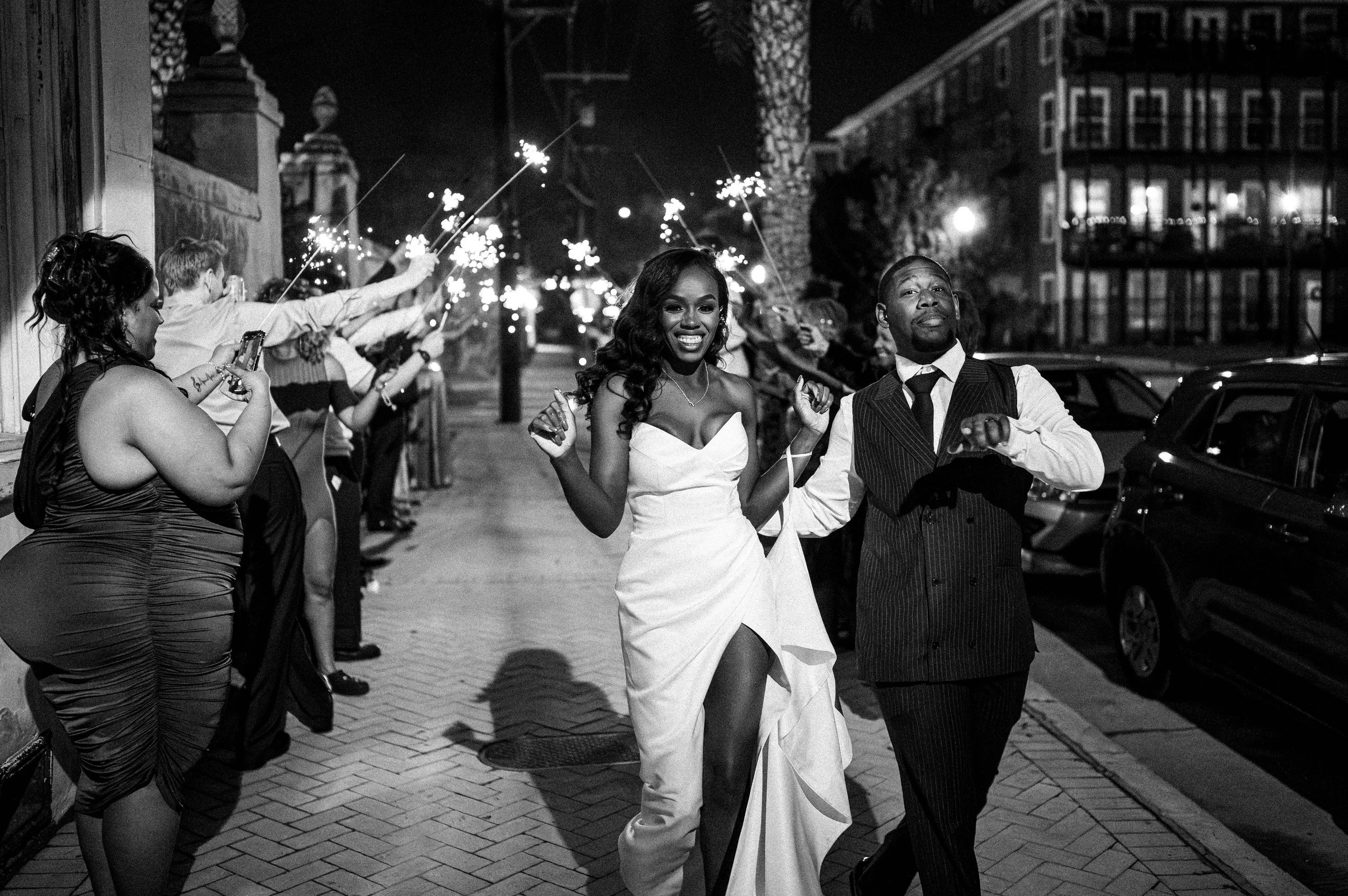 A black and white photo of a woman in a wedding dress and a man in a vest and tie walking together, surrounded by people holding sparklers at night on a New Orleans street.