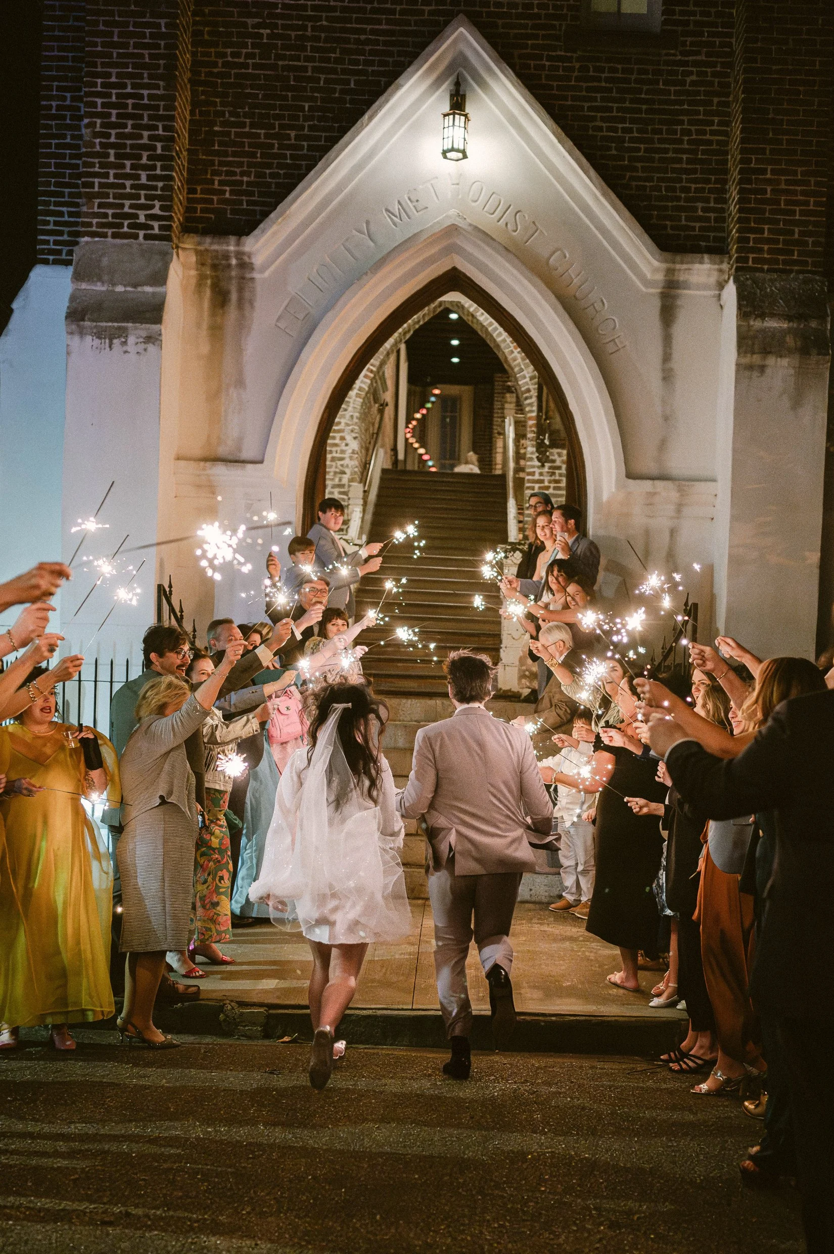 People gather on steps outside a New Orleans church at night, celebrating with sparklers, wedding bride and groom among guests.