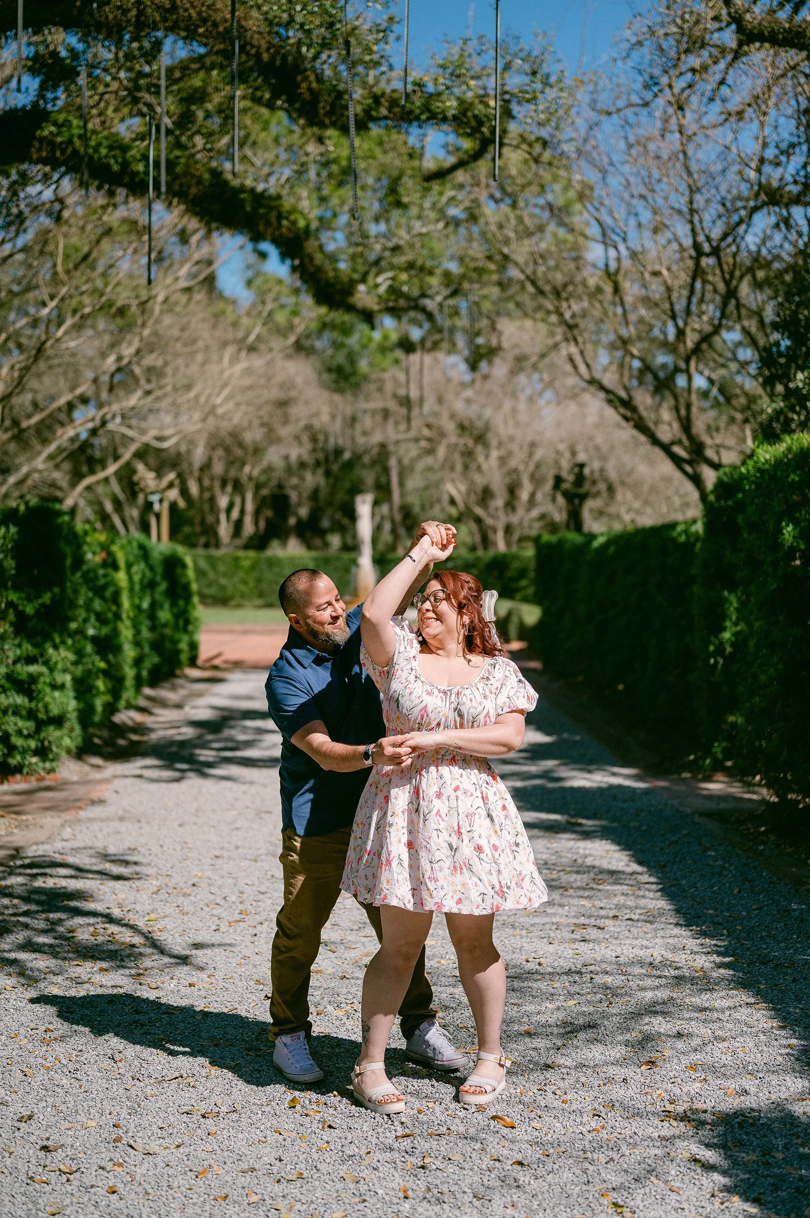 An engaged couple dancing outdoors under a tree with hanging wind chimes on a sunny day.