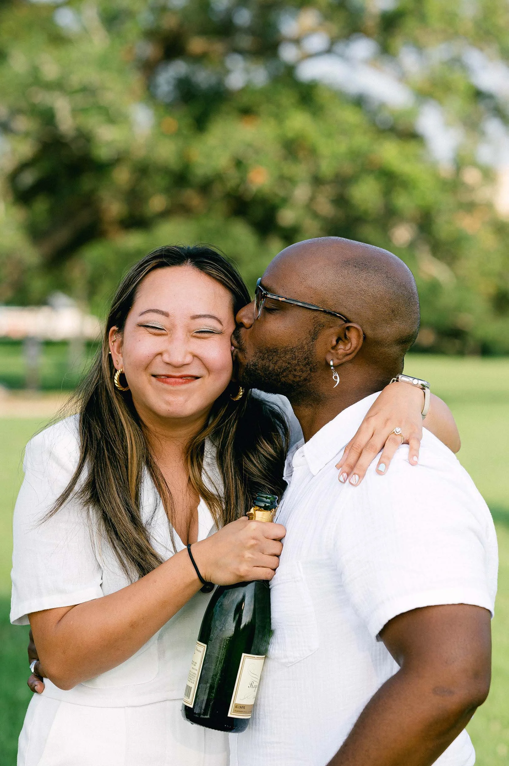 An Asian woman being kissed on the cheek by a black man after being proposed to and saying yes.