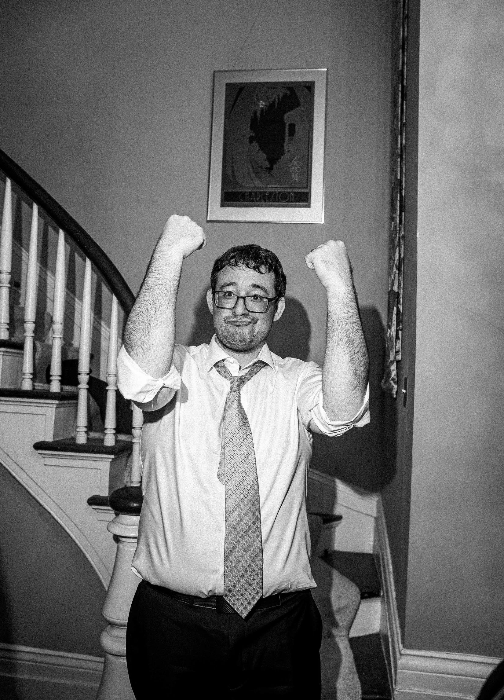 A groom in a dress shirt, tie, and glasses is flexing his biceps and smiling at the camera in celebration of his newlywed status.