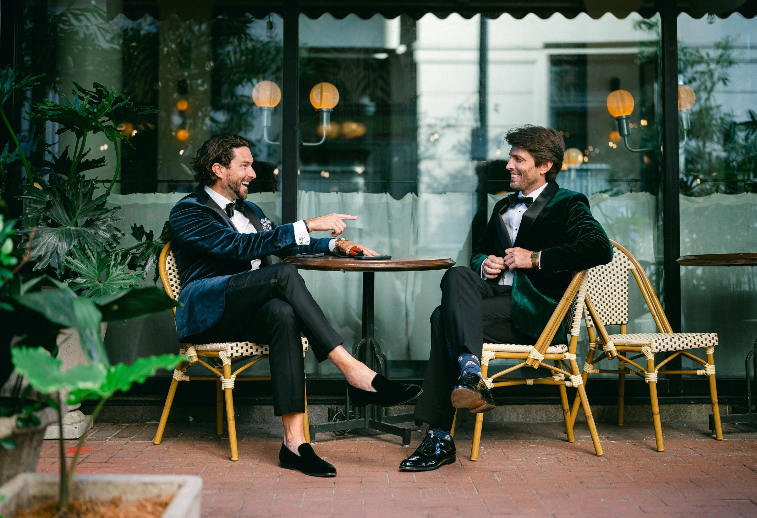 Two grooms in tuxedos sitting at an outdoor cafe table, smiling and laughing, with one pointing at the other. 