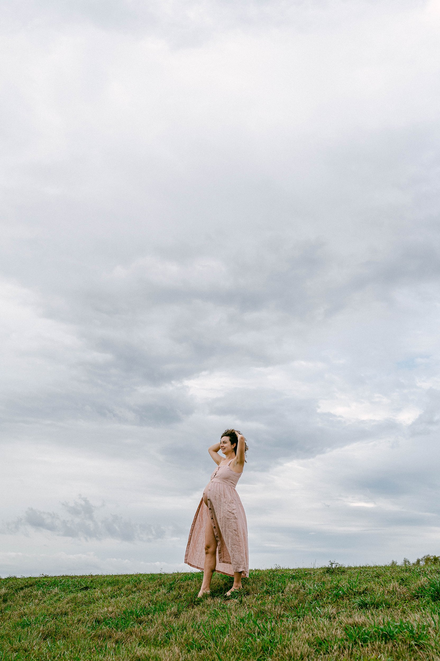 A pregnant woman wearing a pink dress standing on a grassy levee under a cloudy sky, smiling with her hands behind her head.