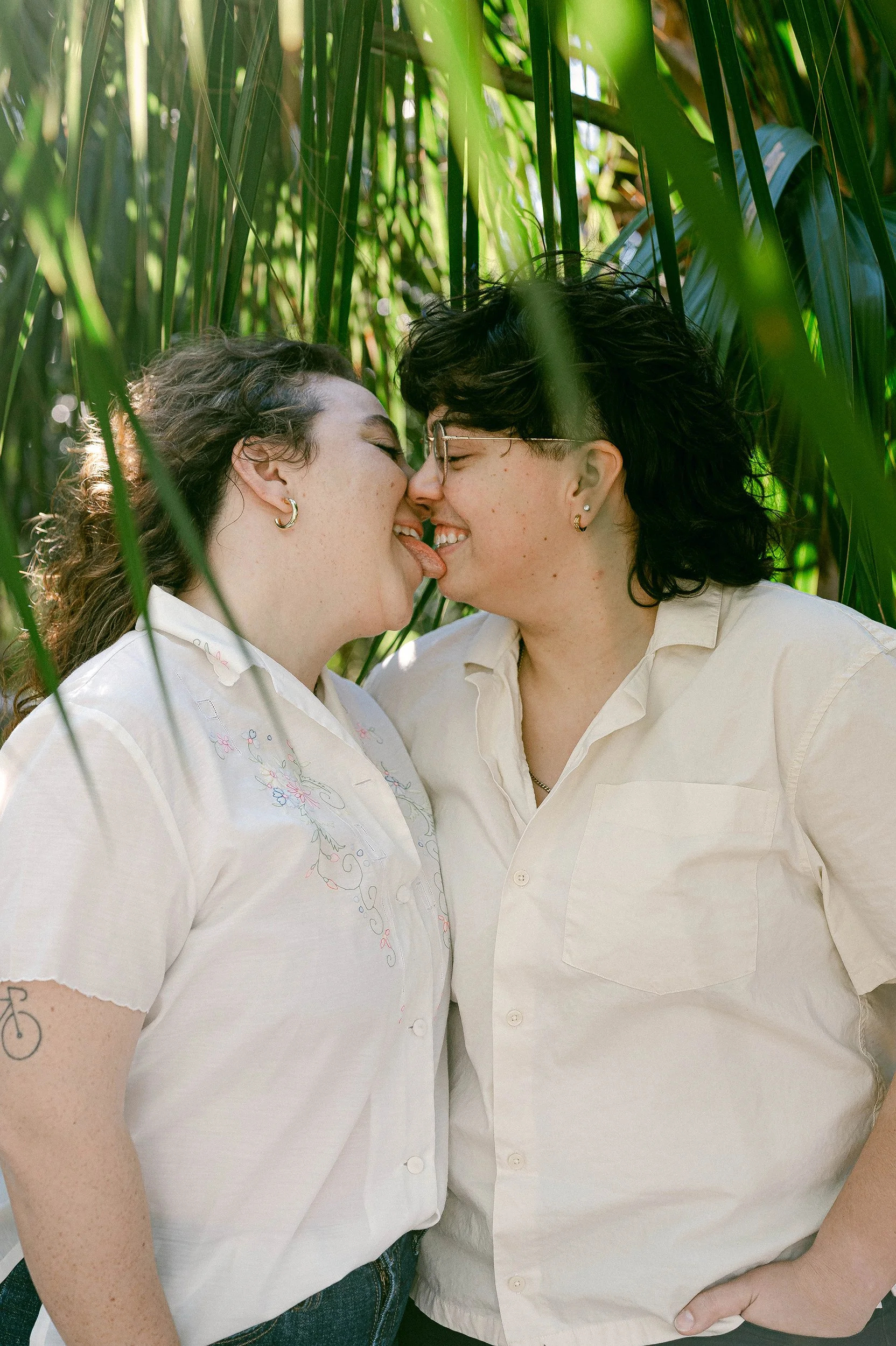 A queer, engaged couple are touching noses and smiling, set against tall green grass and leafy plants.