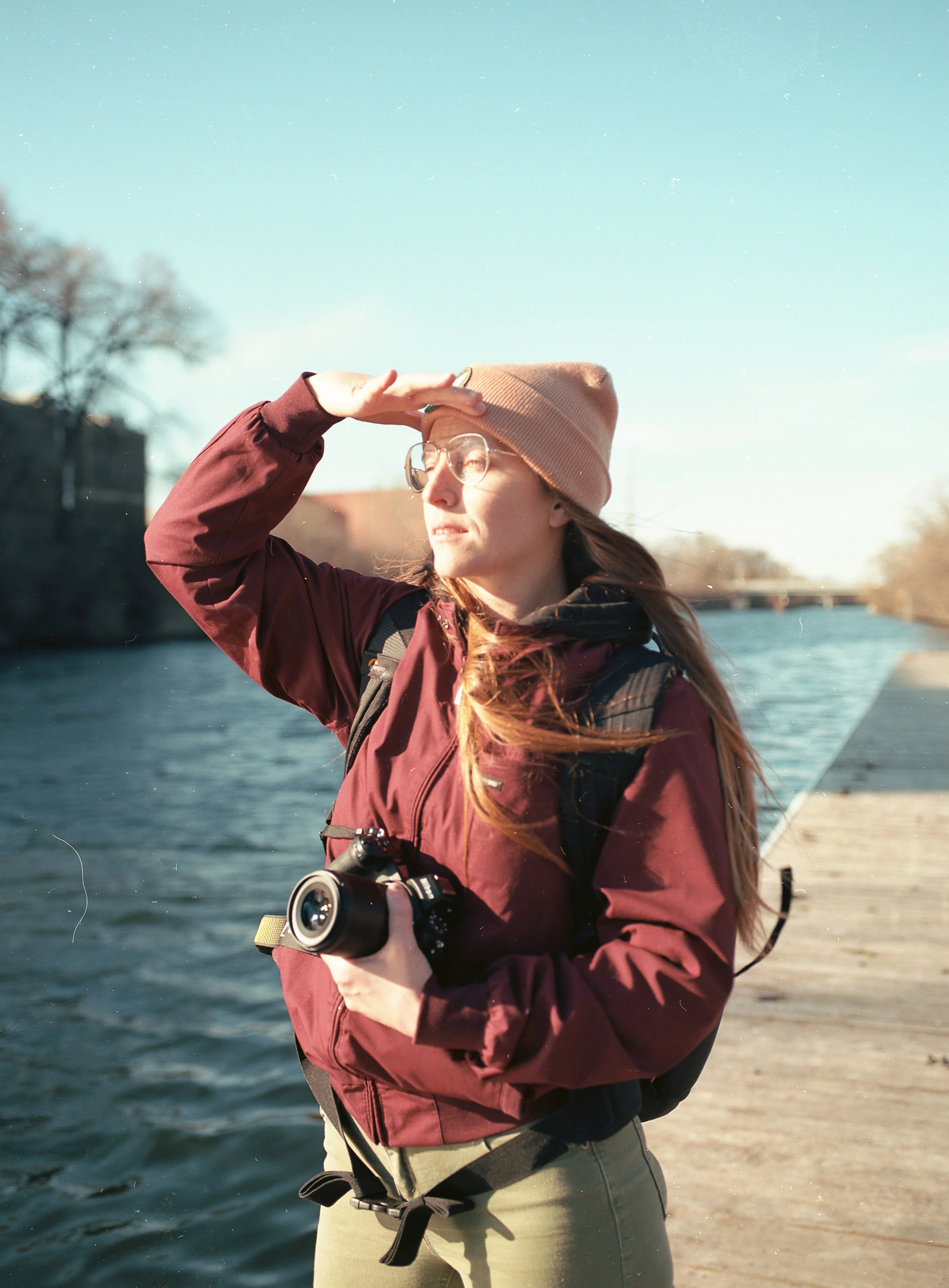 A young woman with long hair, wearing glasses, a tan beanie, and a maroon jacket, standing outdoors near water with trees in the background. She is holding a camera and shielding her eyes from the sun with her hand.