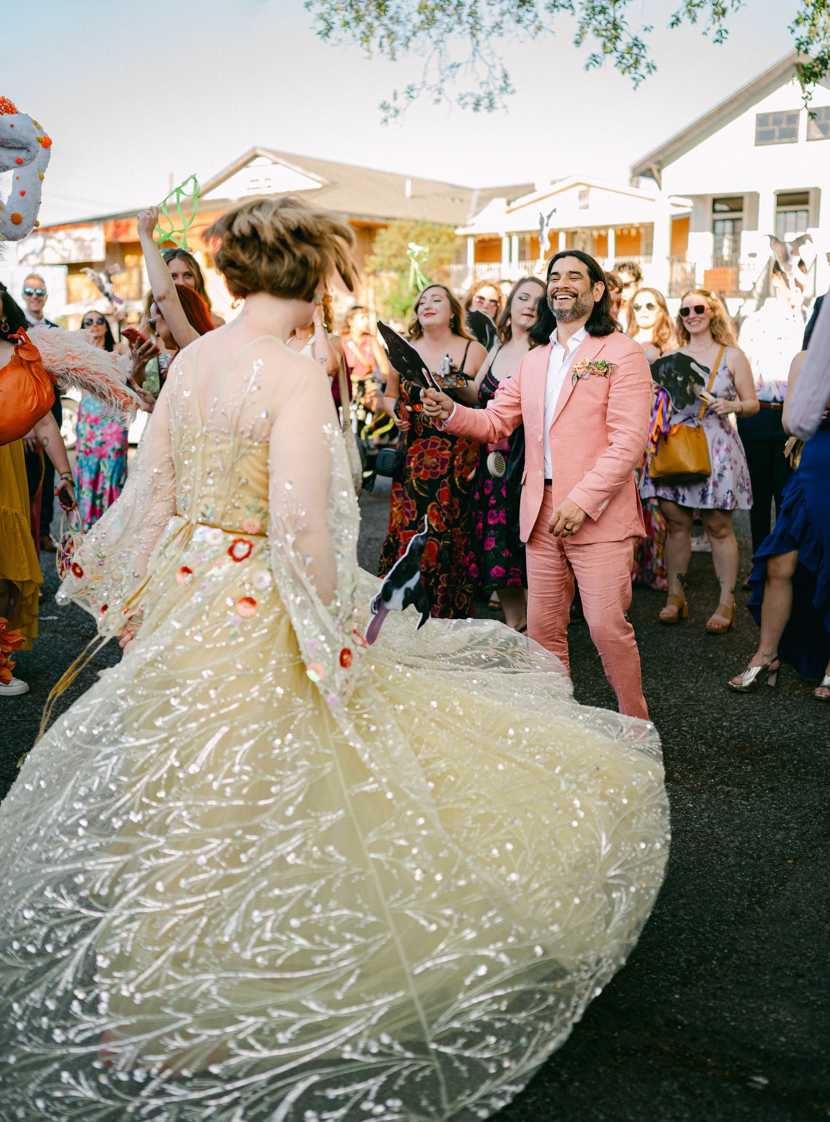 A wedding celebration outdoors with a bride in a voluminous yellow dress and a groom in a pink suit surrounded by guests.