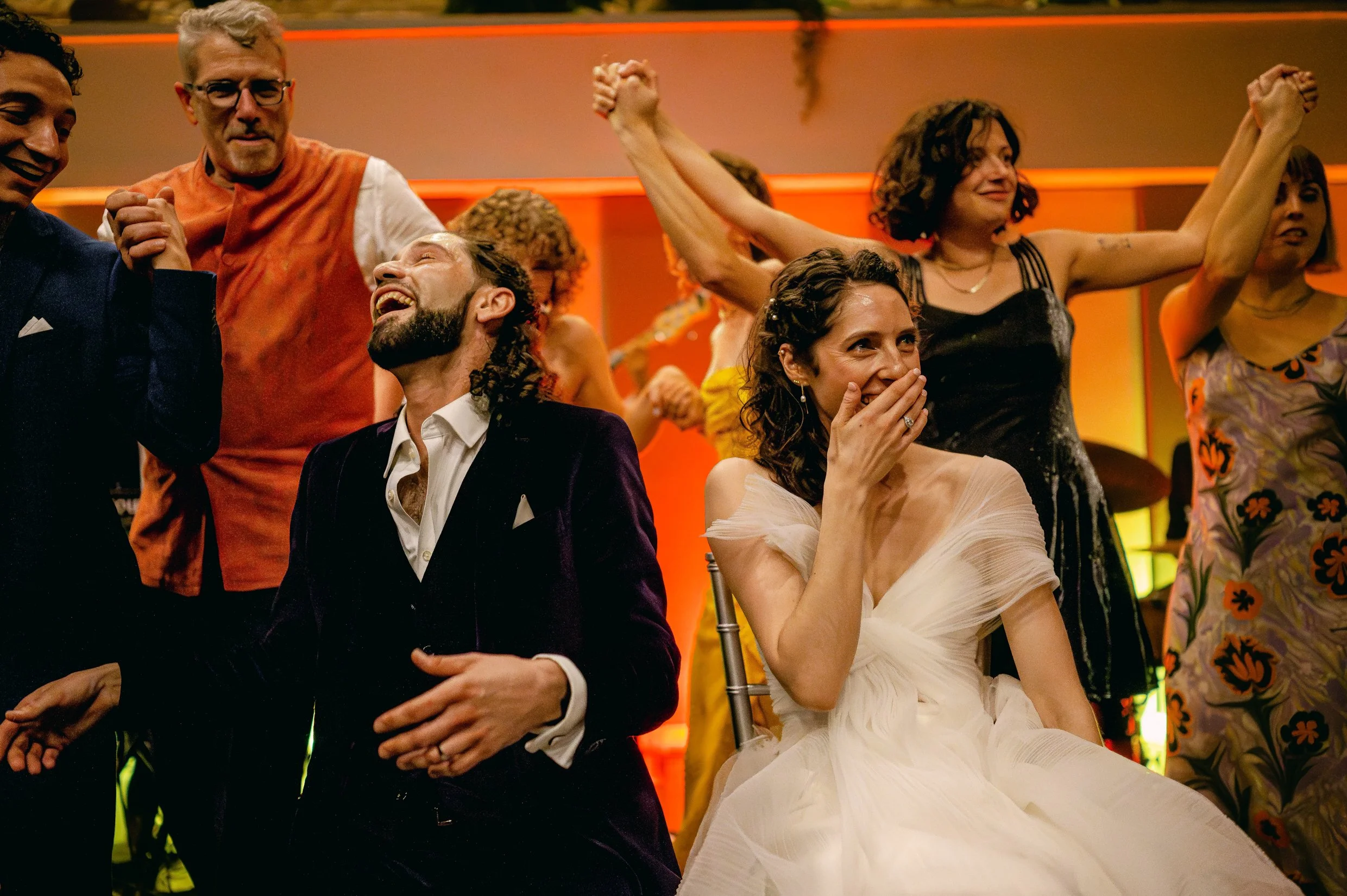 A group of people celebrating during a wedding hora. The bride and groom smile and laugh while guests dance around them. 