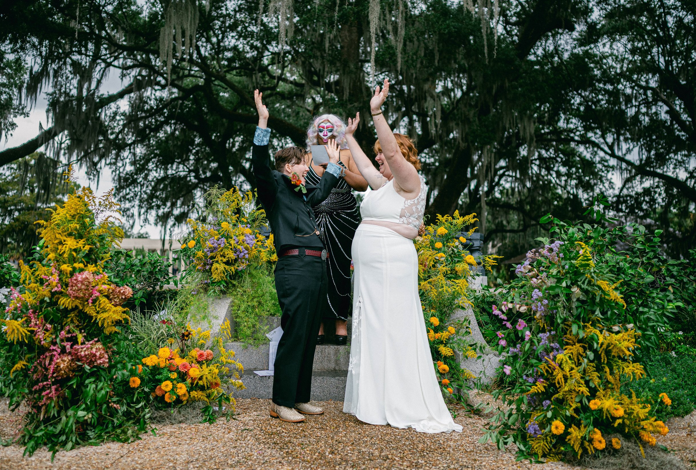 A wedding ceremony with queer people in front of a large tree and surrounded by colorful flowers and have their hands thrown into the air in celebration.