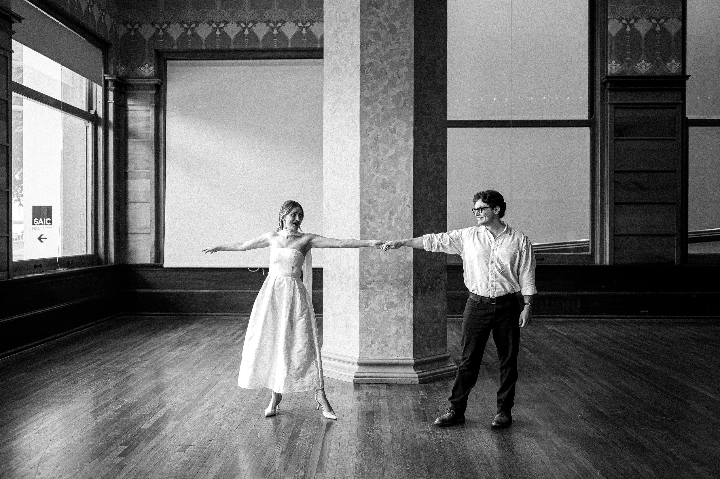 A woman in a bridal dress holding hands with a man in casual clothing, both smiling, inside a room with large windows and wooden floors.