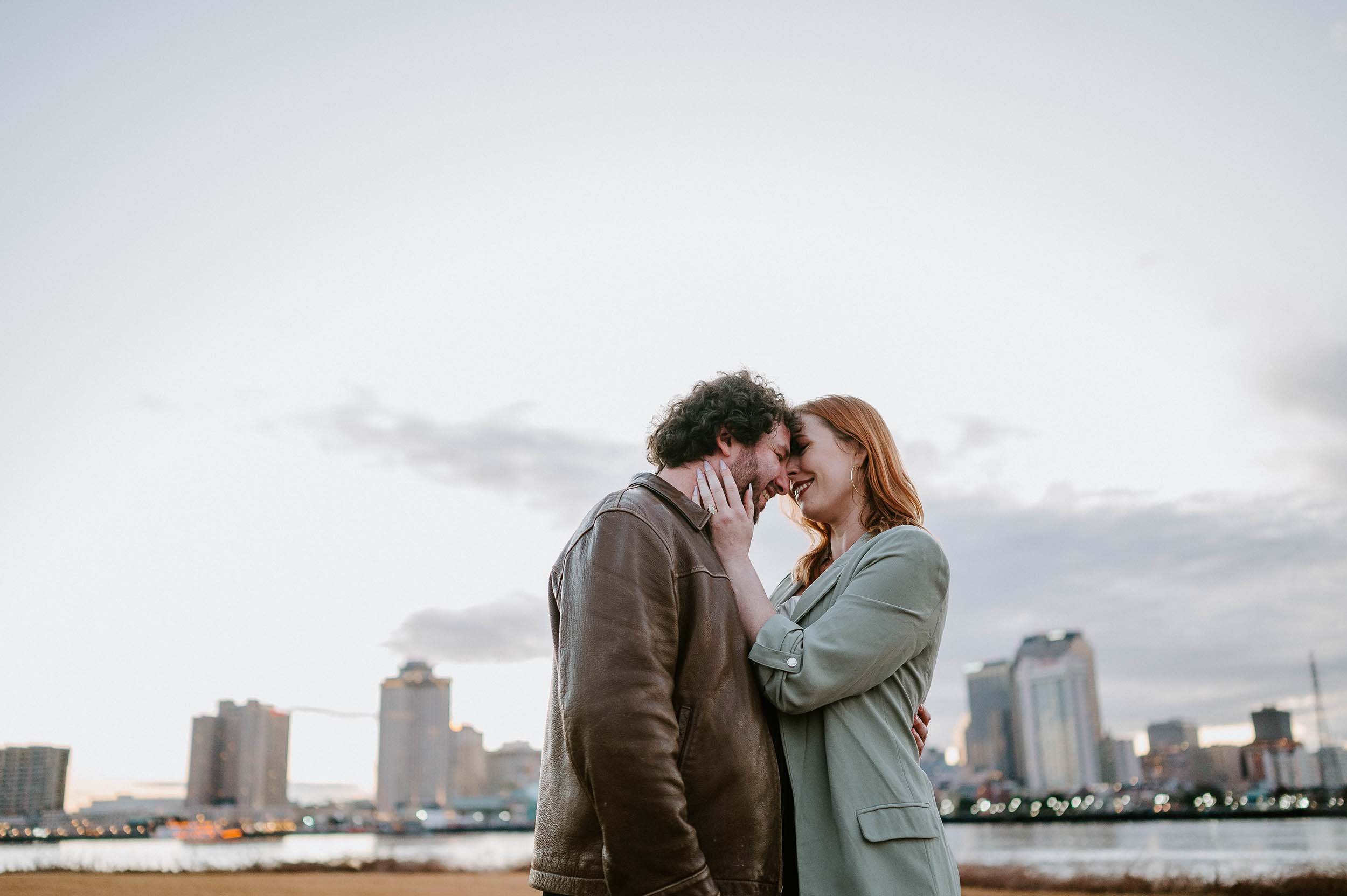 A man and woman are touching foreheads and smiling, standing outdoors with the New Orleans skyline in the background during dusk.