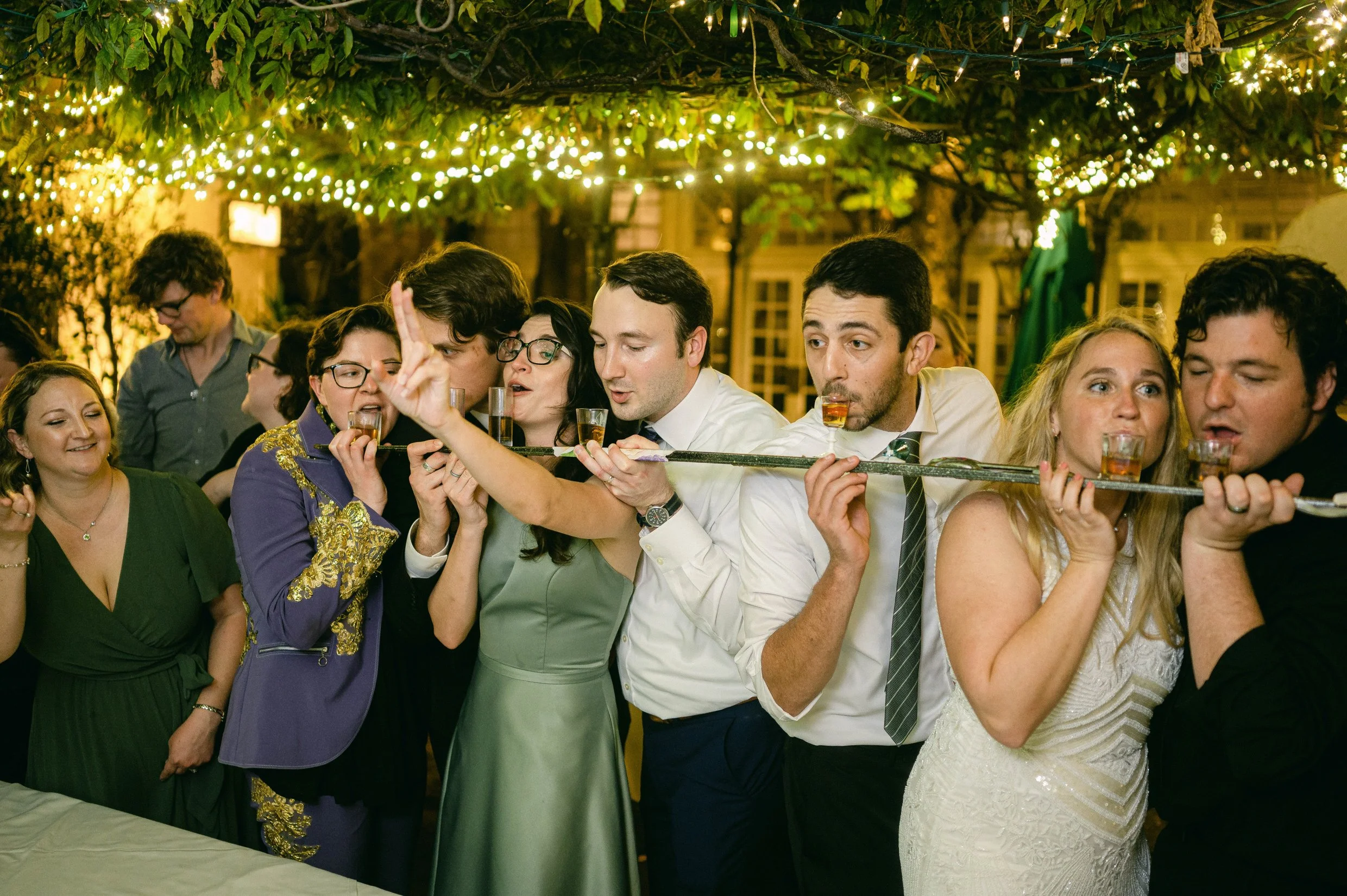 A group of people celebrating at a wedding, sharing a shot-ski under string lights hanging from trees.