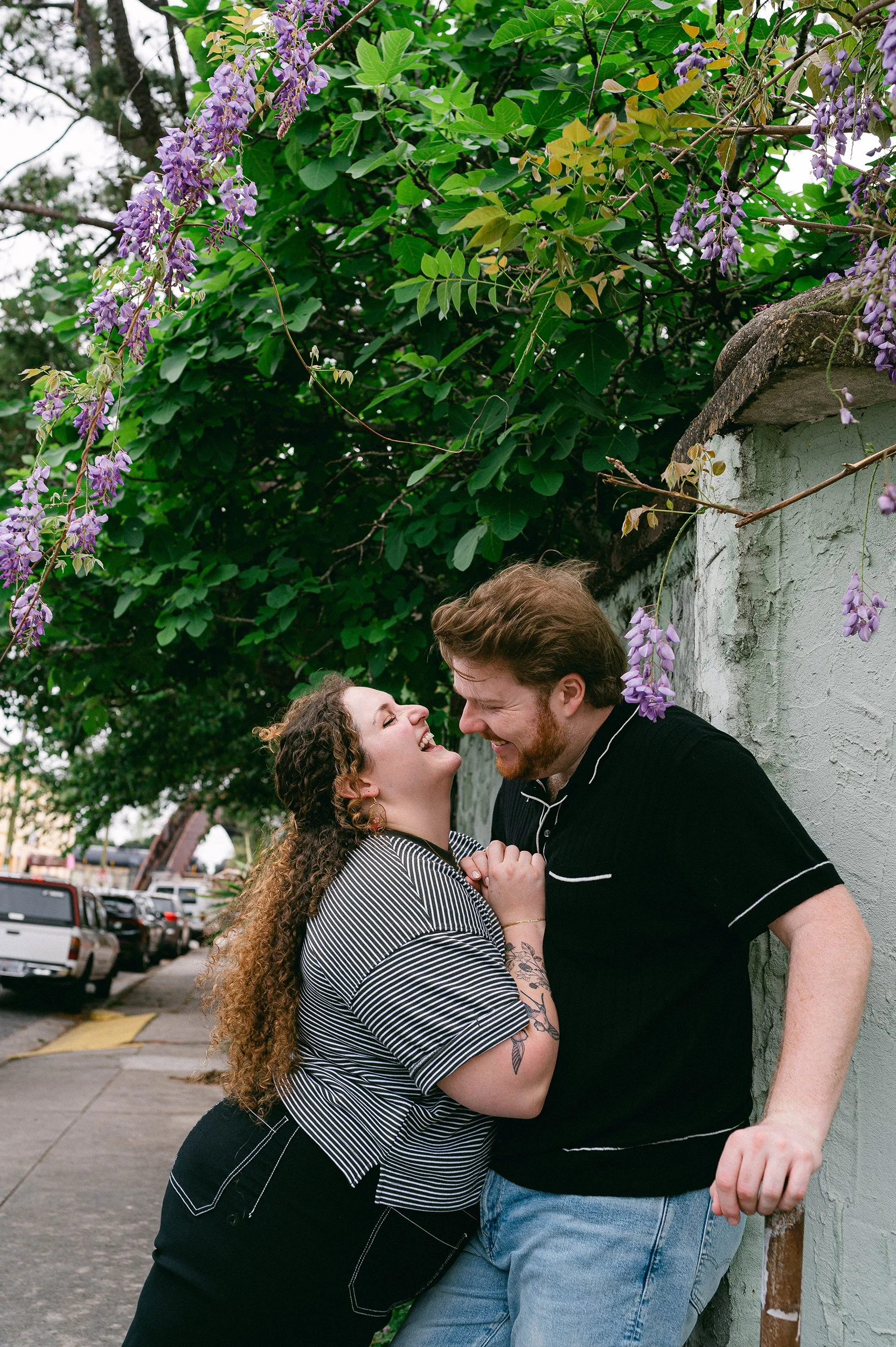 An engaged couple laughs and smiles closely together outdoors during the daytime, with the woman holding the man's hands, against a wall with purple flowering vines. 