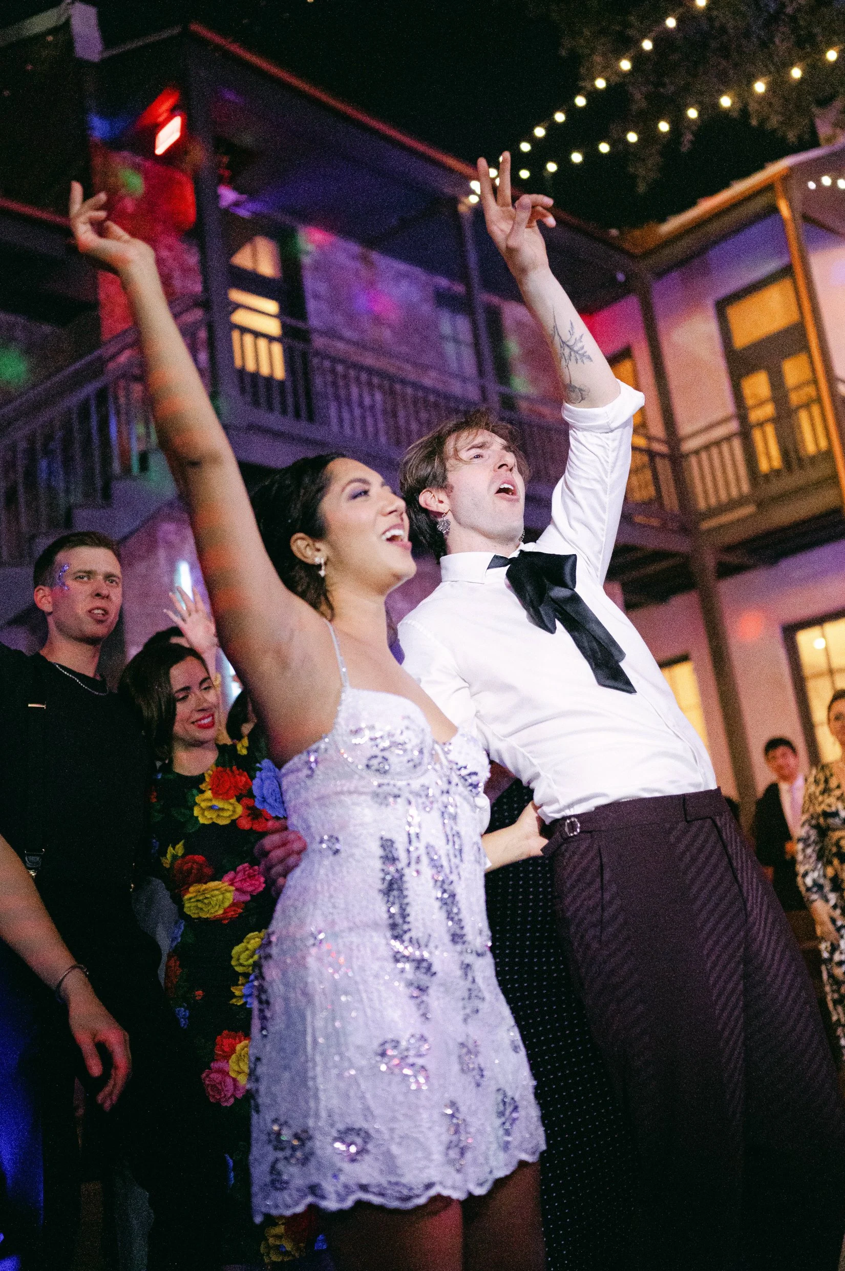 A bride and groom and their guests dancing at a lively indoor party with string lights and a balcony in the background.
