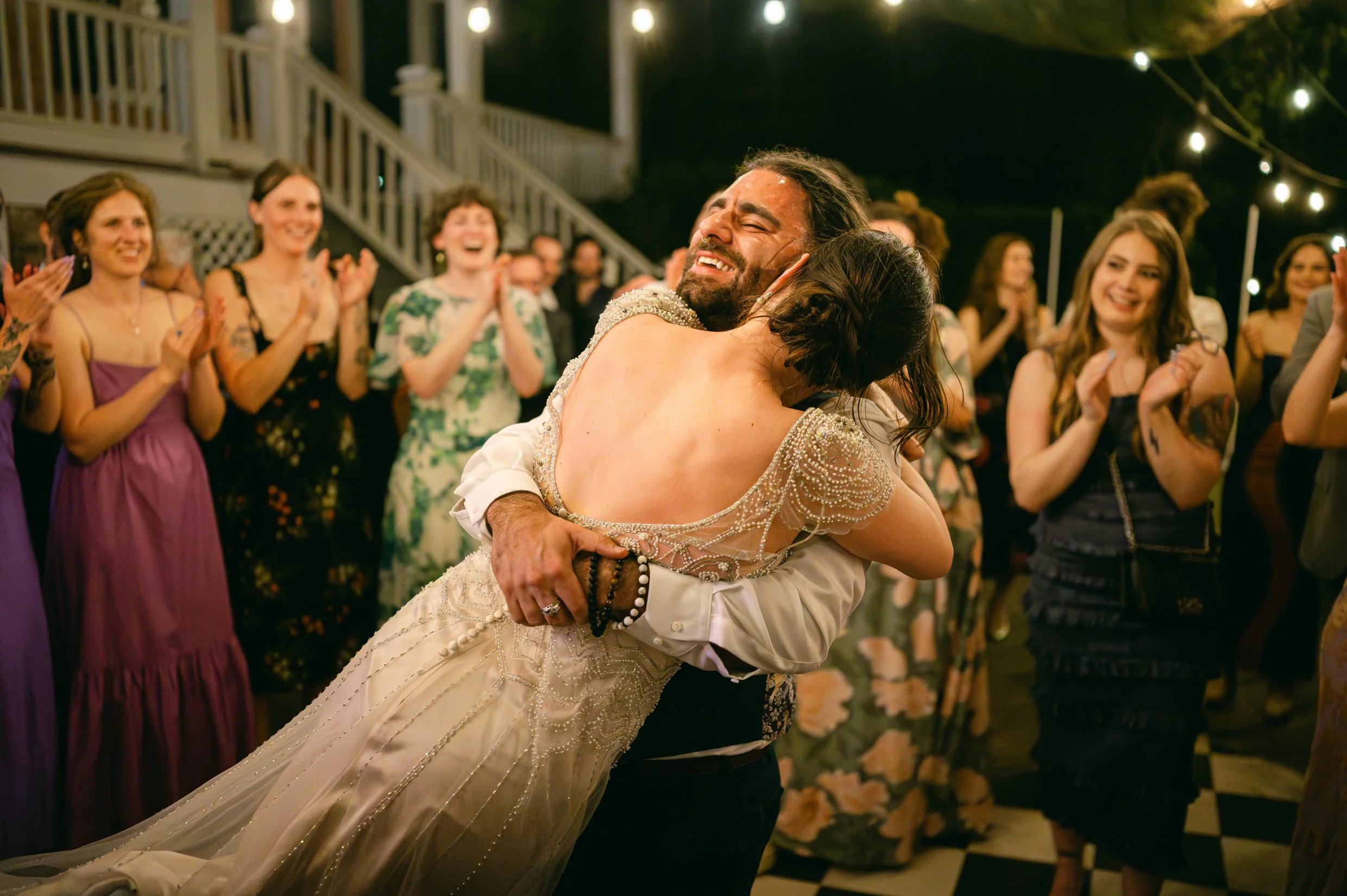 A bride and groom dancing closely at their wedding reception. They are smiling and embracing each other, surrounded by guests clapping and cheering.