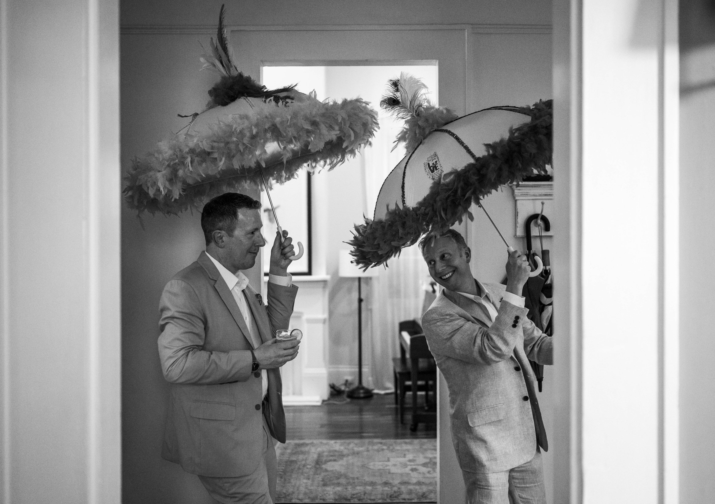 Two grooms dressed in suits holding feathered parasols inside a building, with a woman smiling in the background.